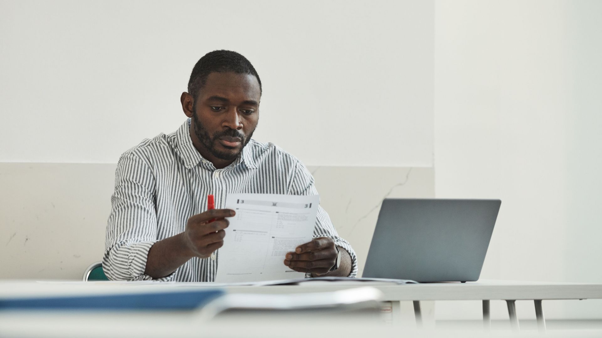 An adult man attentively grading test papers at his workspace with a laptop.