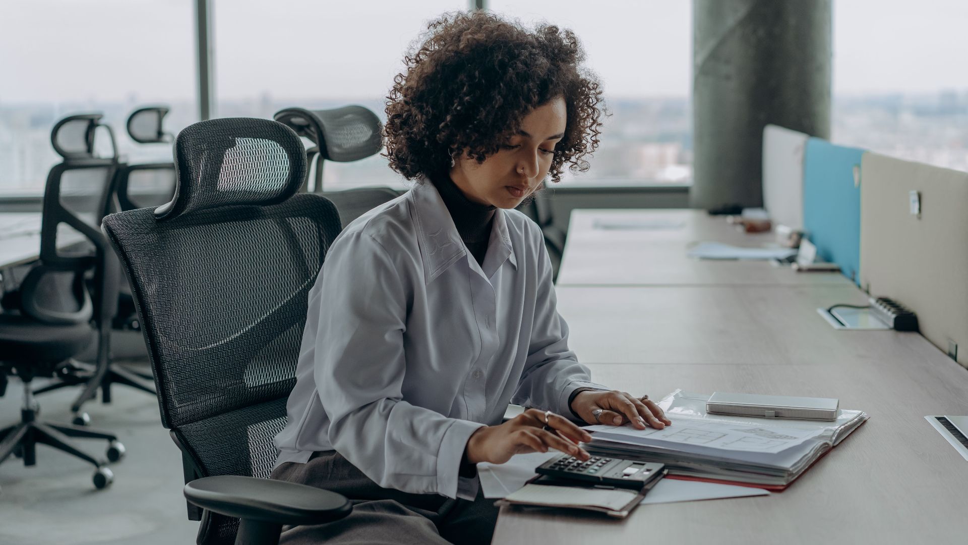 A professional woman with curly hair calculating documents in a modern office setting.