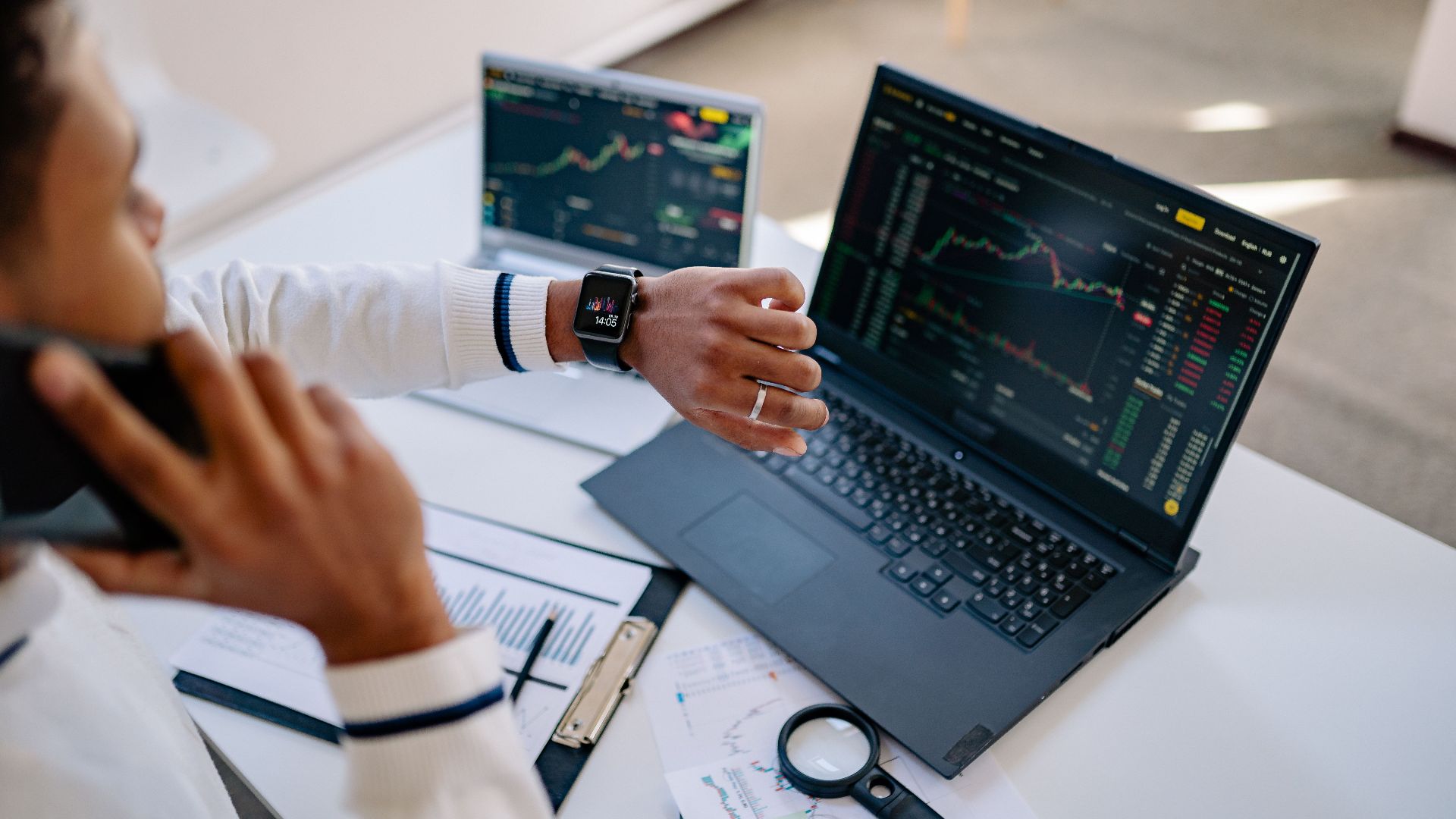 Man multitasking with a smartwatch and laptop analyzing stock market charts at the office.