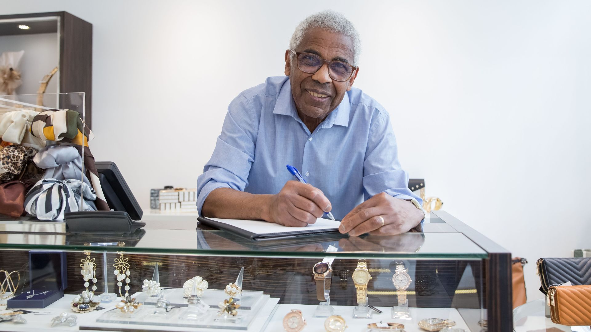 Smiling elderly shopkeeper in Portugal writing at jewelry display counter with eyeglasses.