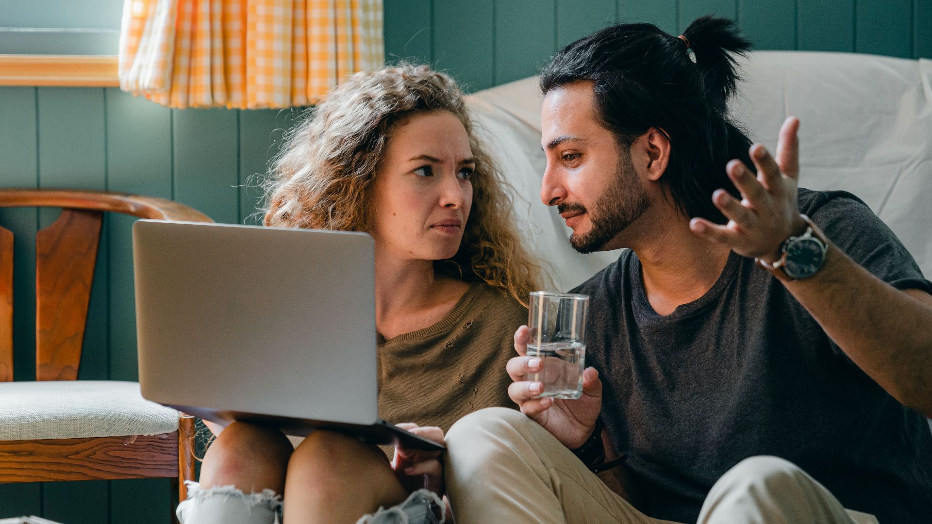 Bewildered young multiethnic couple in casual outfits sitting on floor against blue wall while browsing modern netbook and discussing news