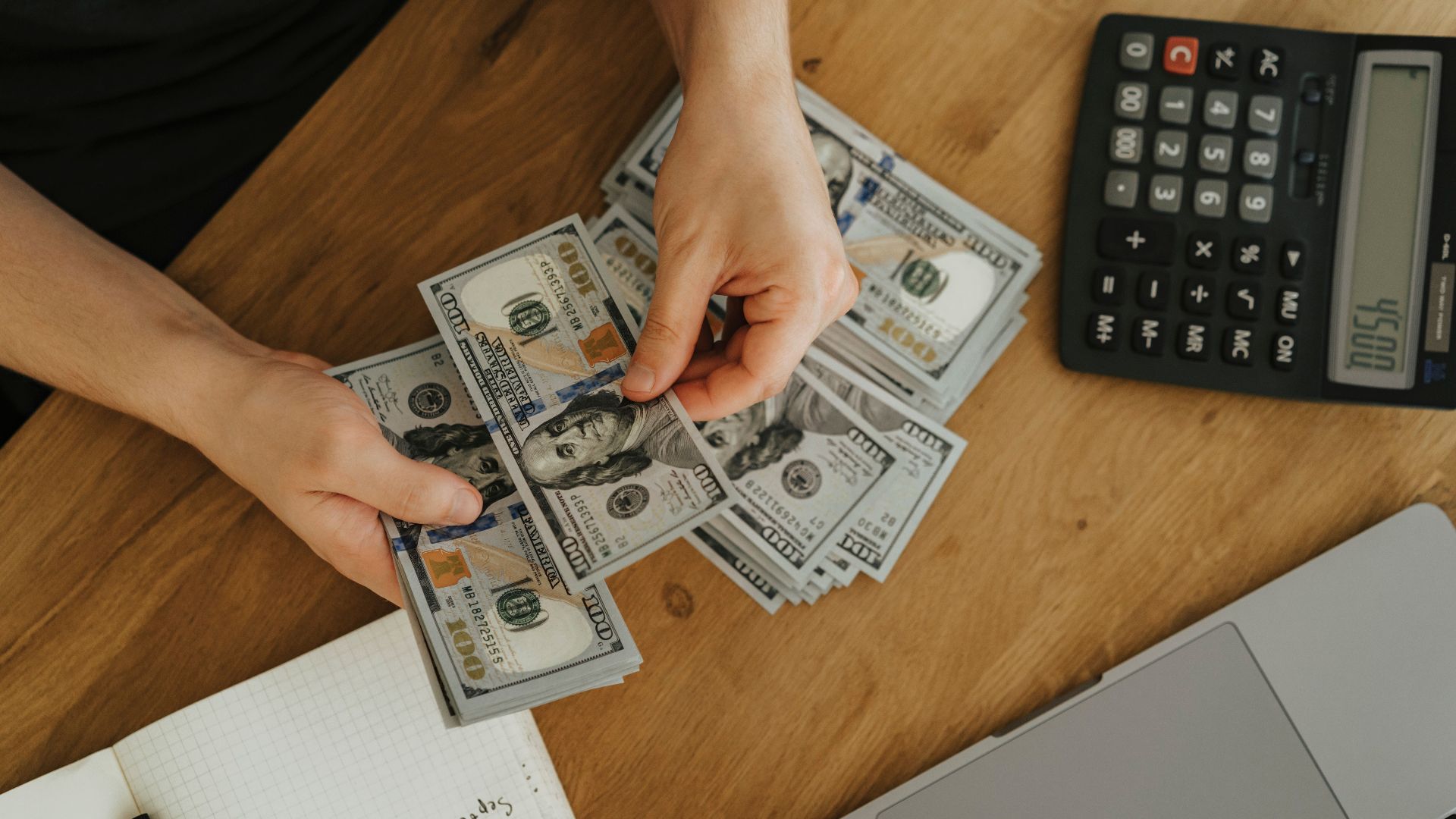 Close-up of hands counting dollar bills with calculator, notebook, and laptop on wooden table.