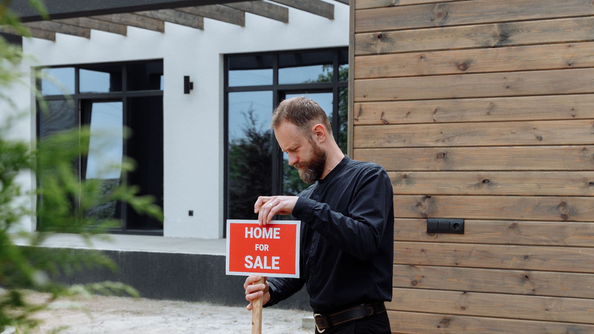 A real estate agent holding a home for sale sign in front of a modern house.