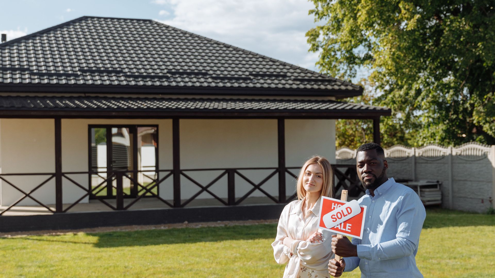 Diverse couple celebrating new home purchase with a sold sign outdoors.