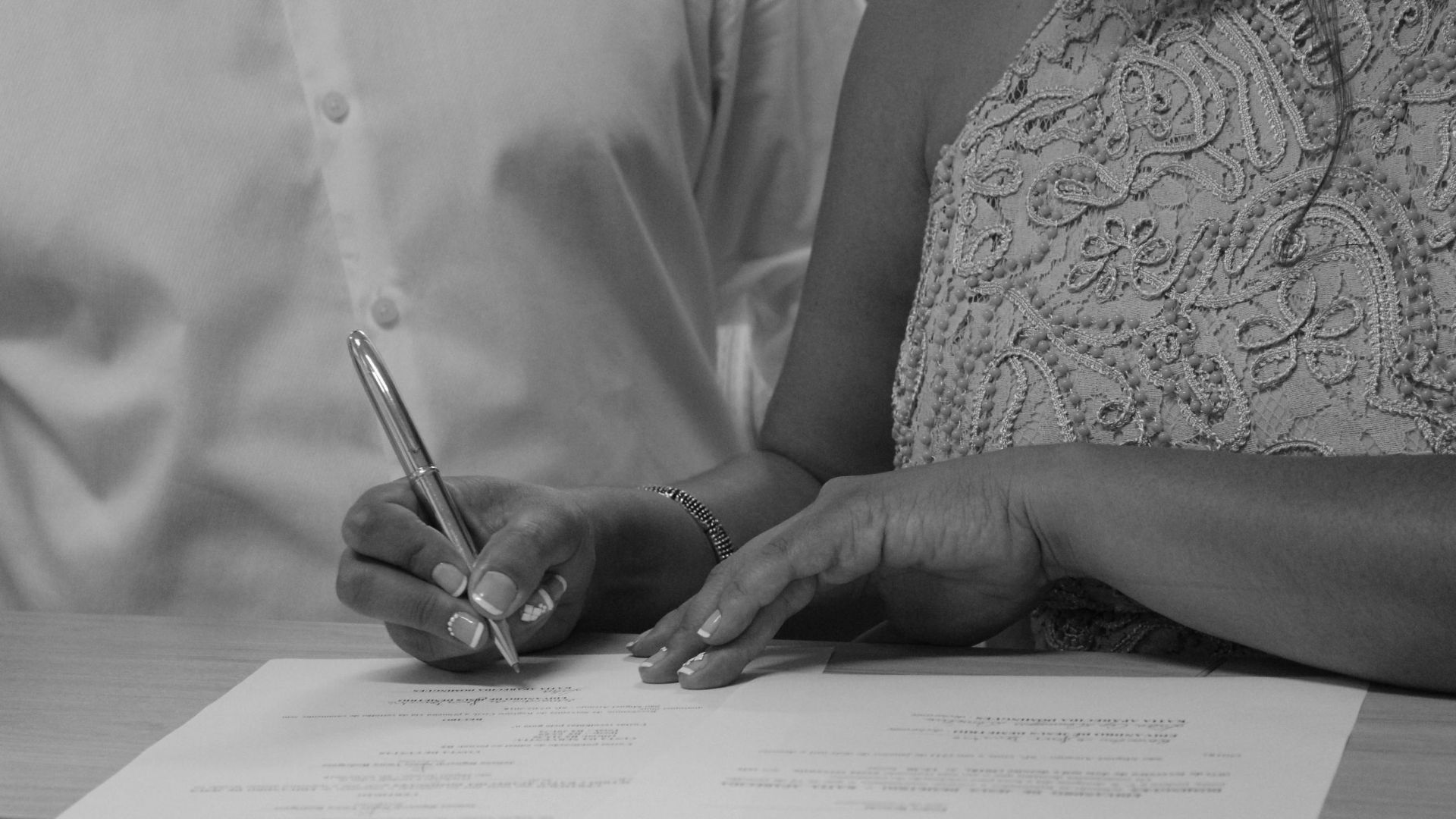 Black and white image of a couple signing documents, focusing on their hands and pen.