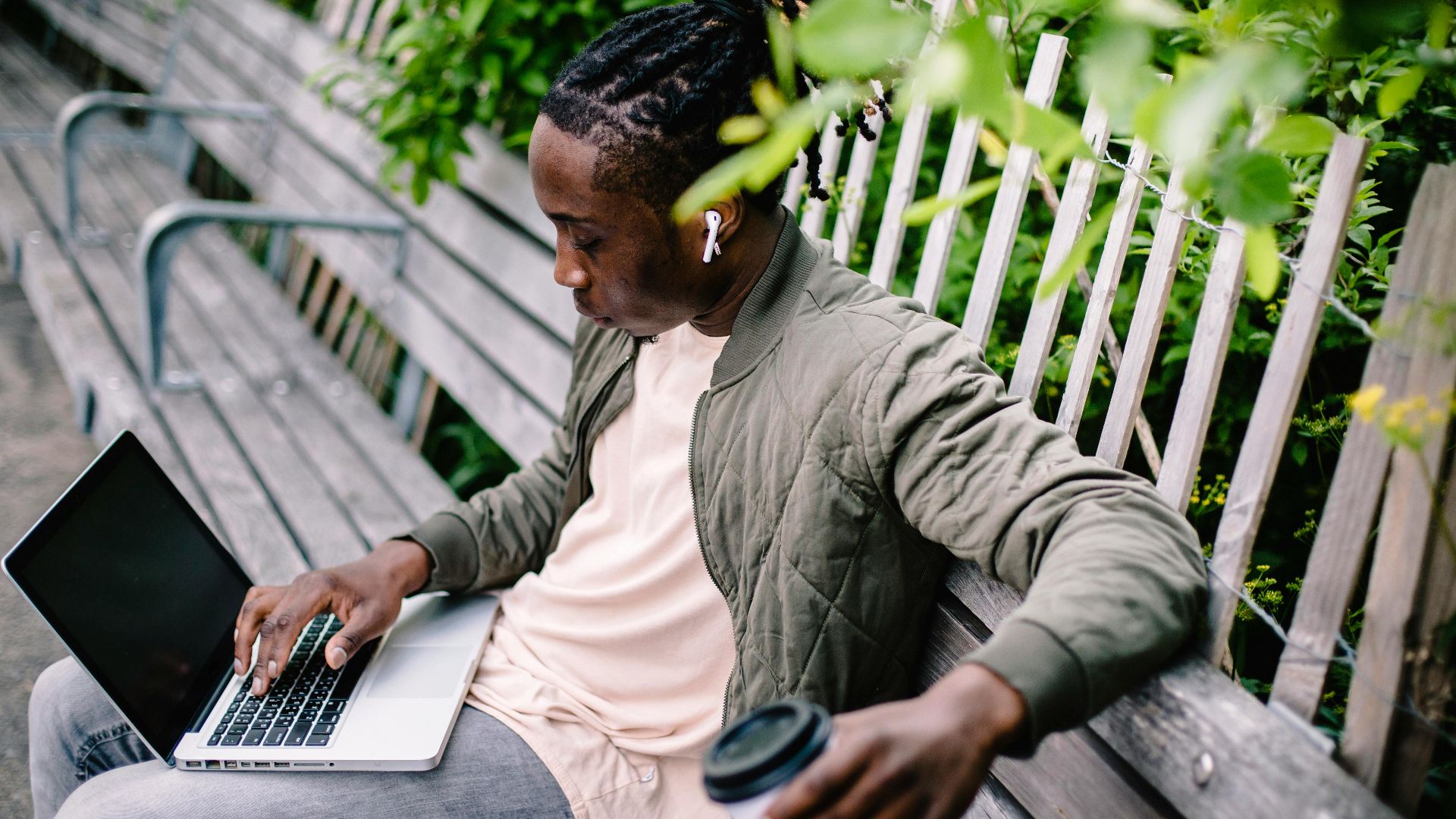 From above of young relaxed African American male freelancer in casual wear with wireless earbuds in ears sitting on bench and working on project on laptop in quiet green park