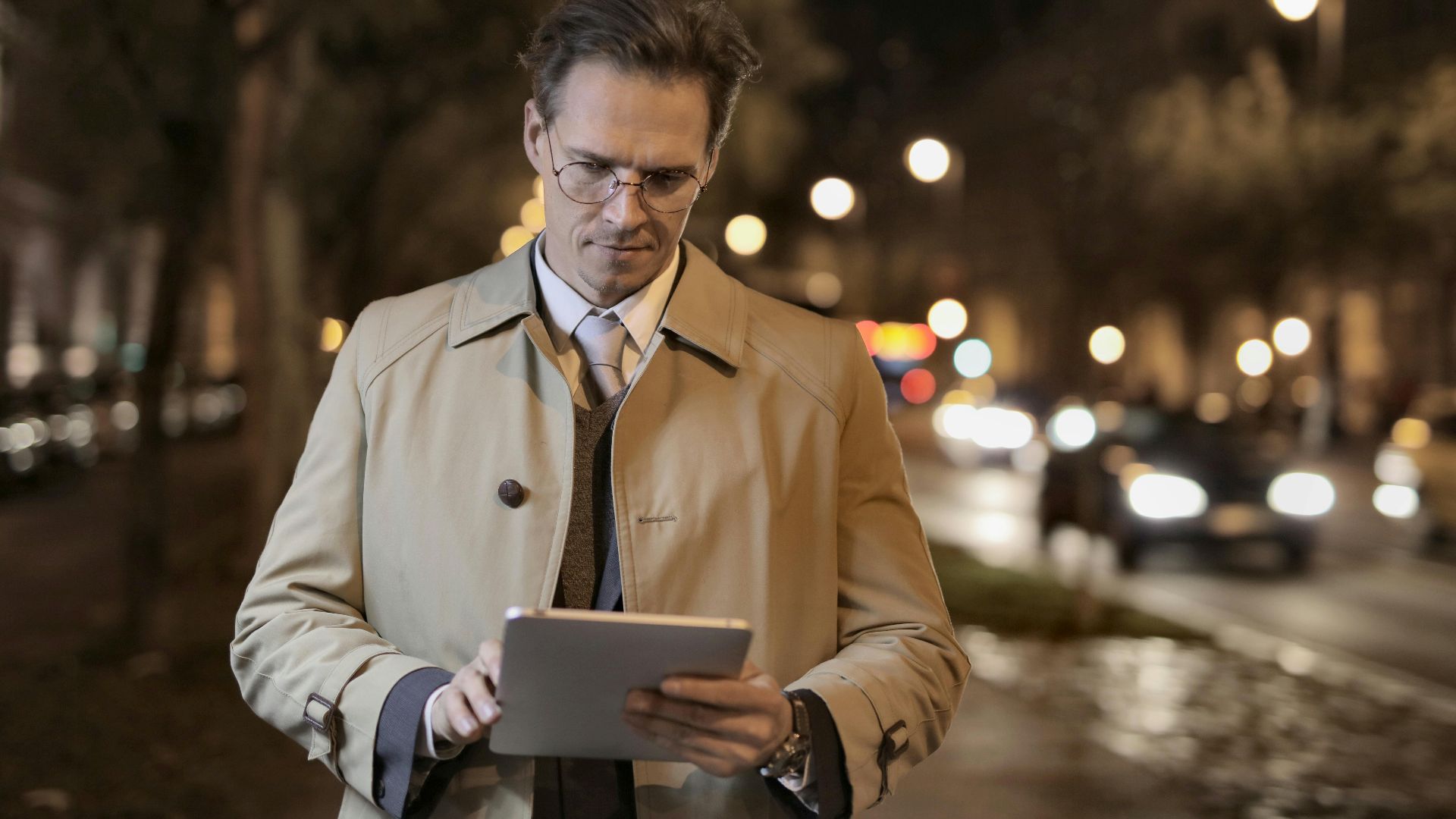 Positive serious male in glasses standing on street near road while surfing internet on tablet