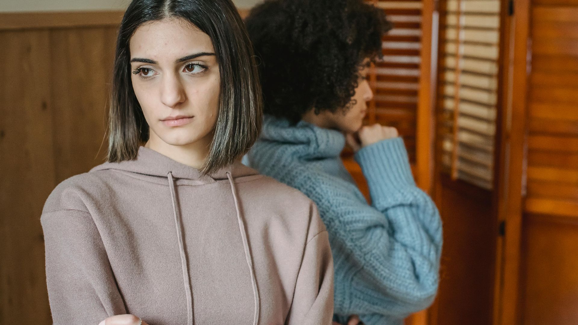 Upset multiracial women in casual wear standing in light room near wall with wooden folding screen while having conflict at home