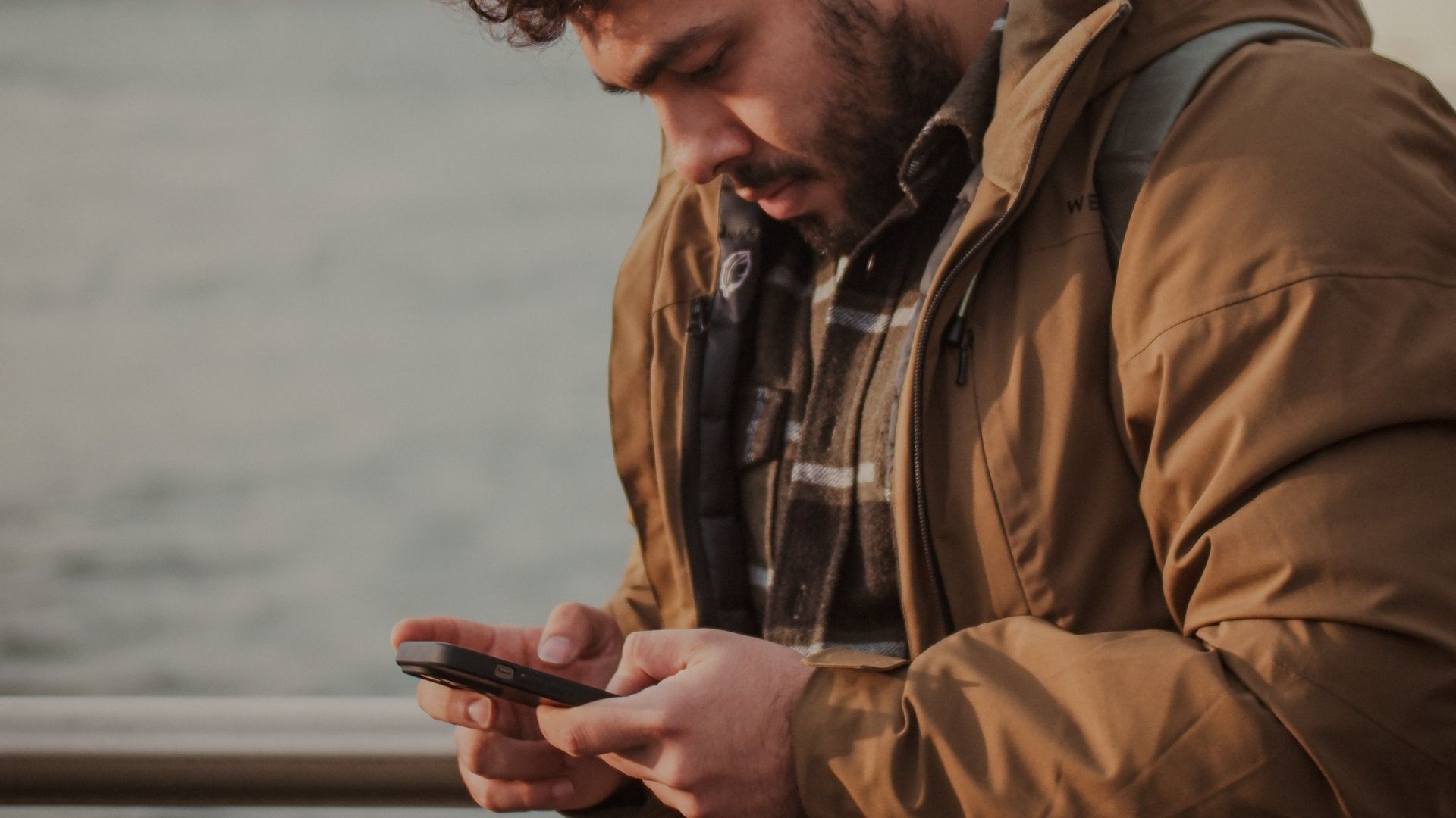 Man wearing a jacket, checking his smartphone by a waterfront, cityscape in the background.