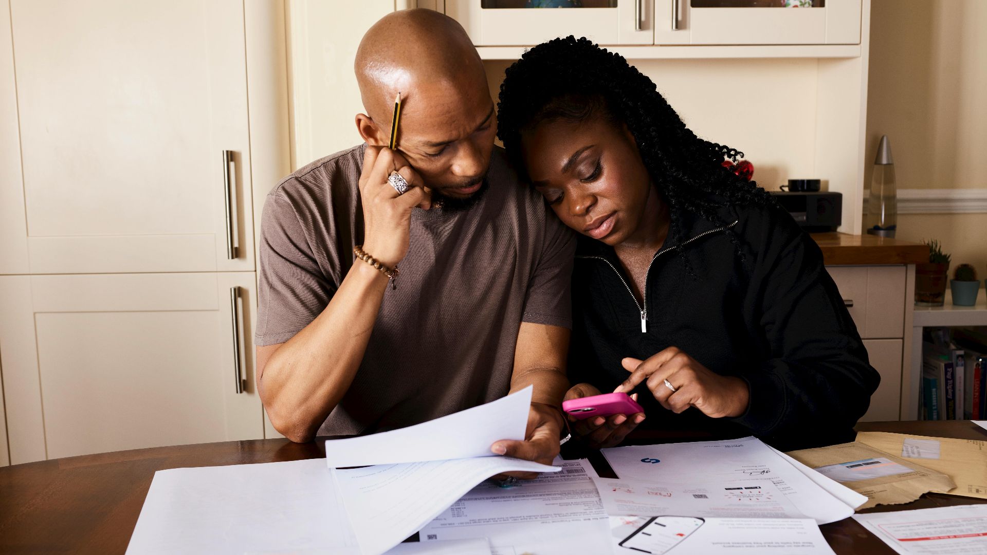 A couple sits at a table managing domestic finances, evaluating documents and using a smartphone.