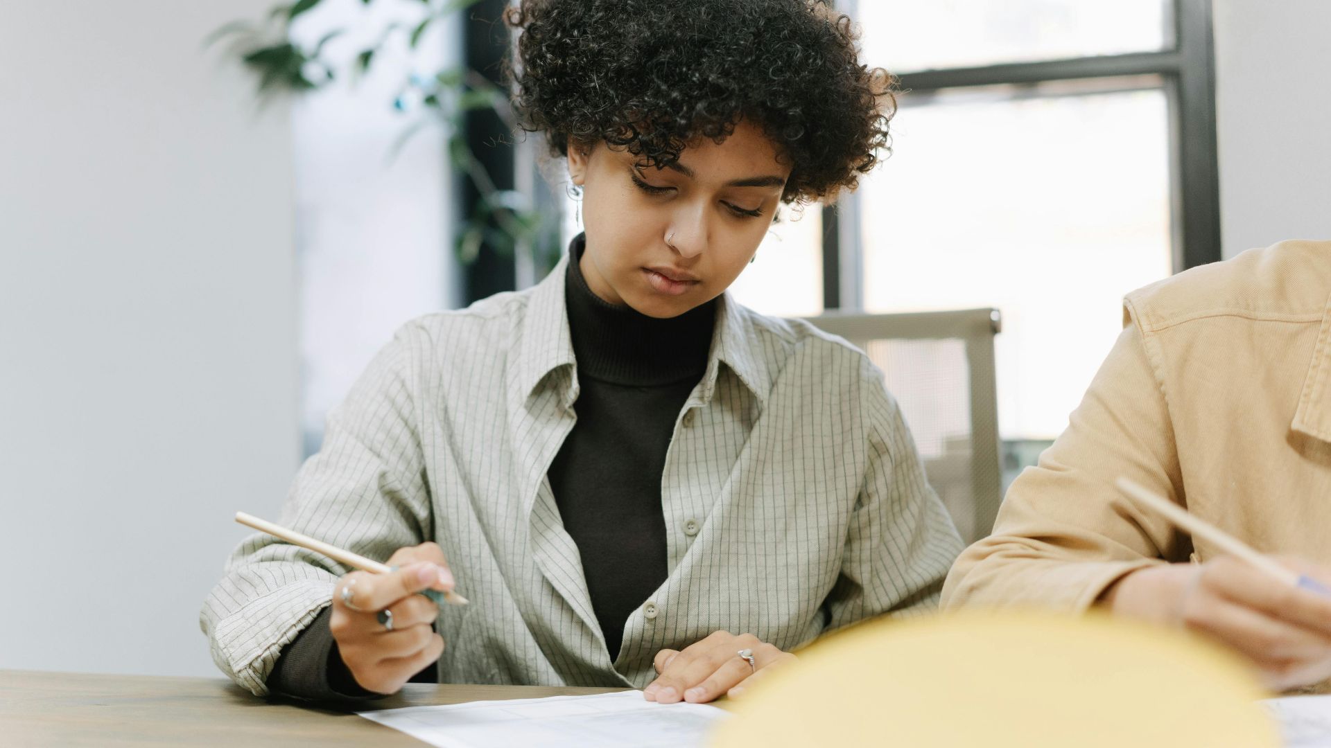 A young woman with curly hair focused on writing with a pen at a desk indoors.