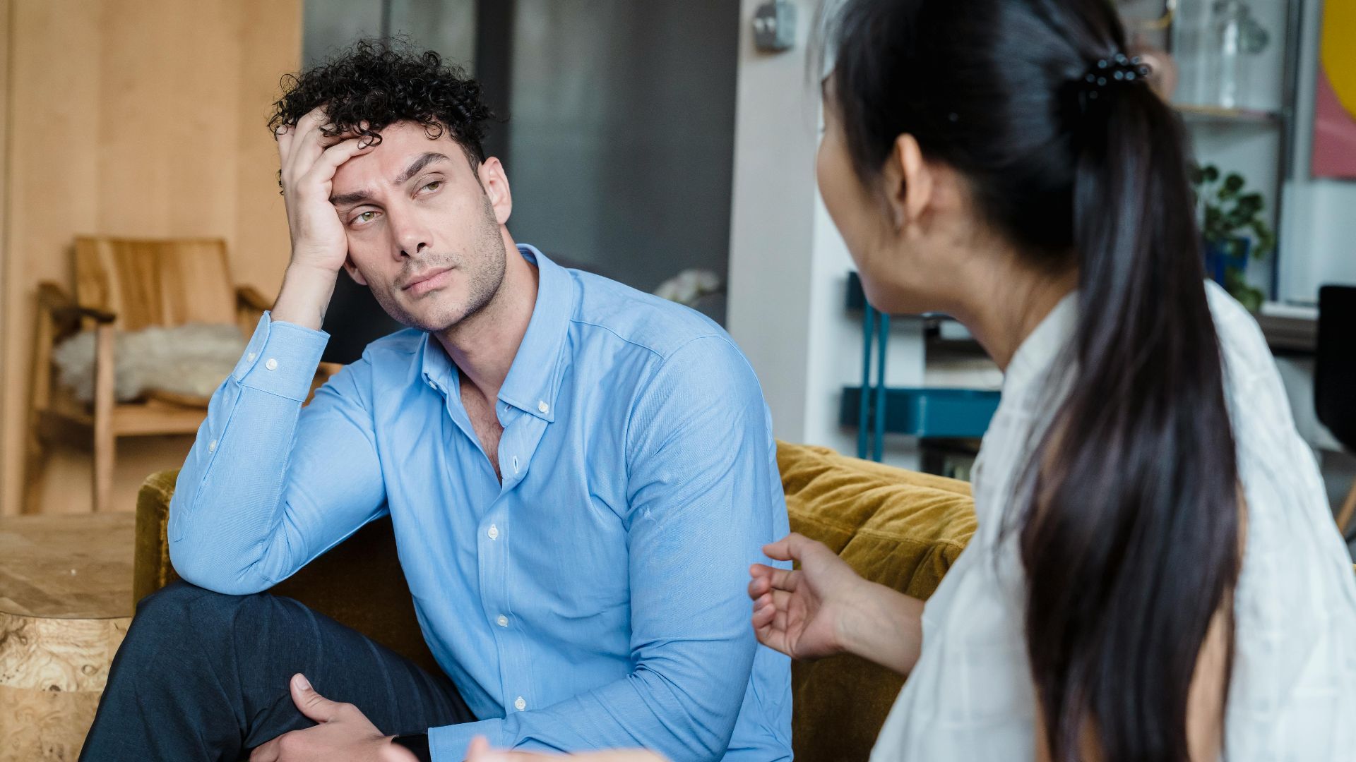 A tired man in a blue shirt listens while a woman talks indoors. Modern living room.