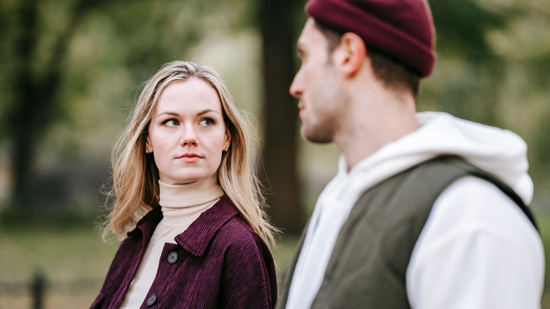 Calm woman in casual warm clothes with long hair looking at boyfriend with beard in hat while walking in park