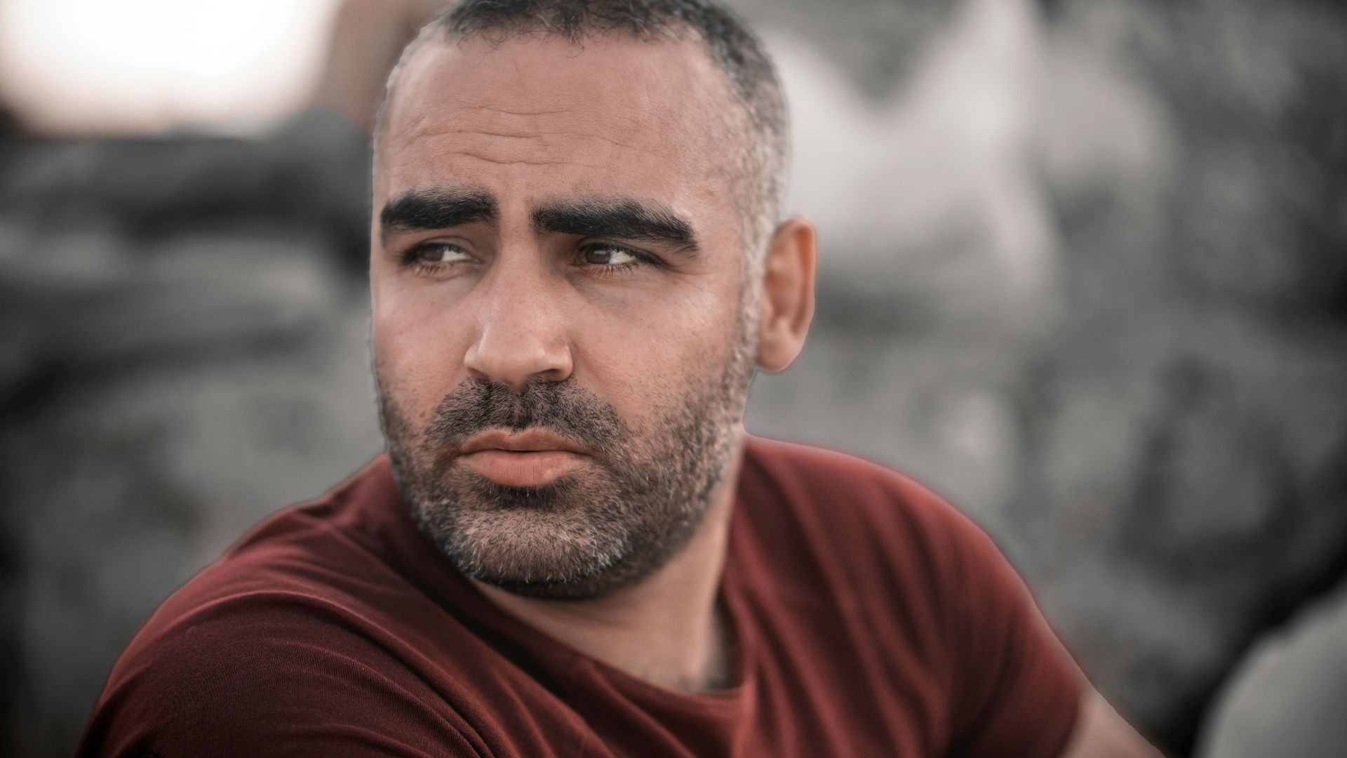 A bearded man wearing a red shirt looks to the side, set against a blurred outdoor backdrop.