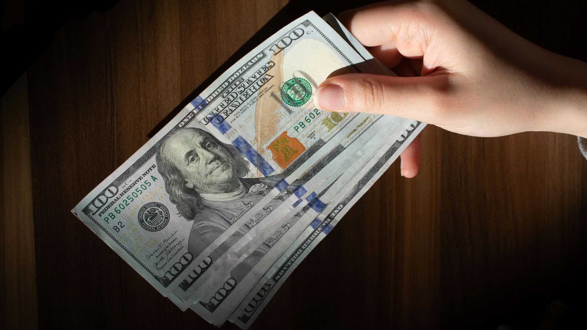 A hand holding a stack of 100 dollar bills on a wooden table, highlighting currency wealth.