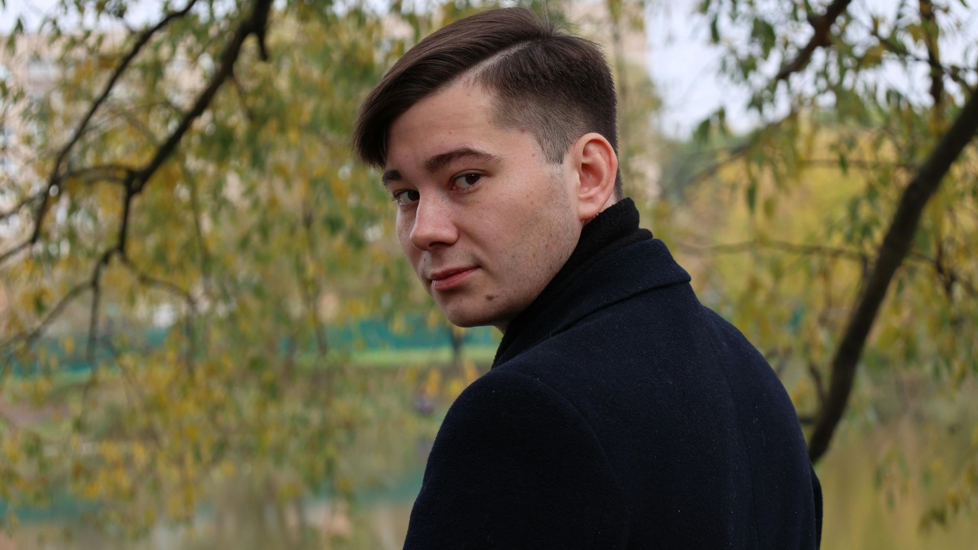 A man in a black coat glances back while standing in a serene autumn park.