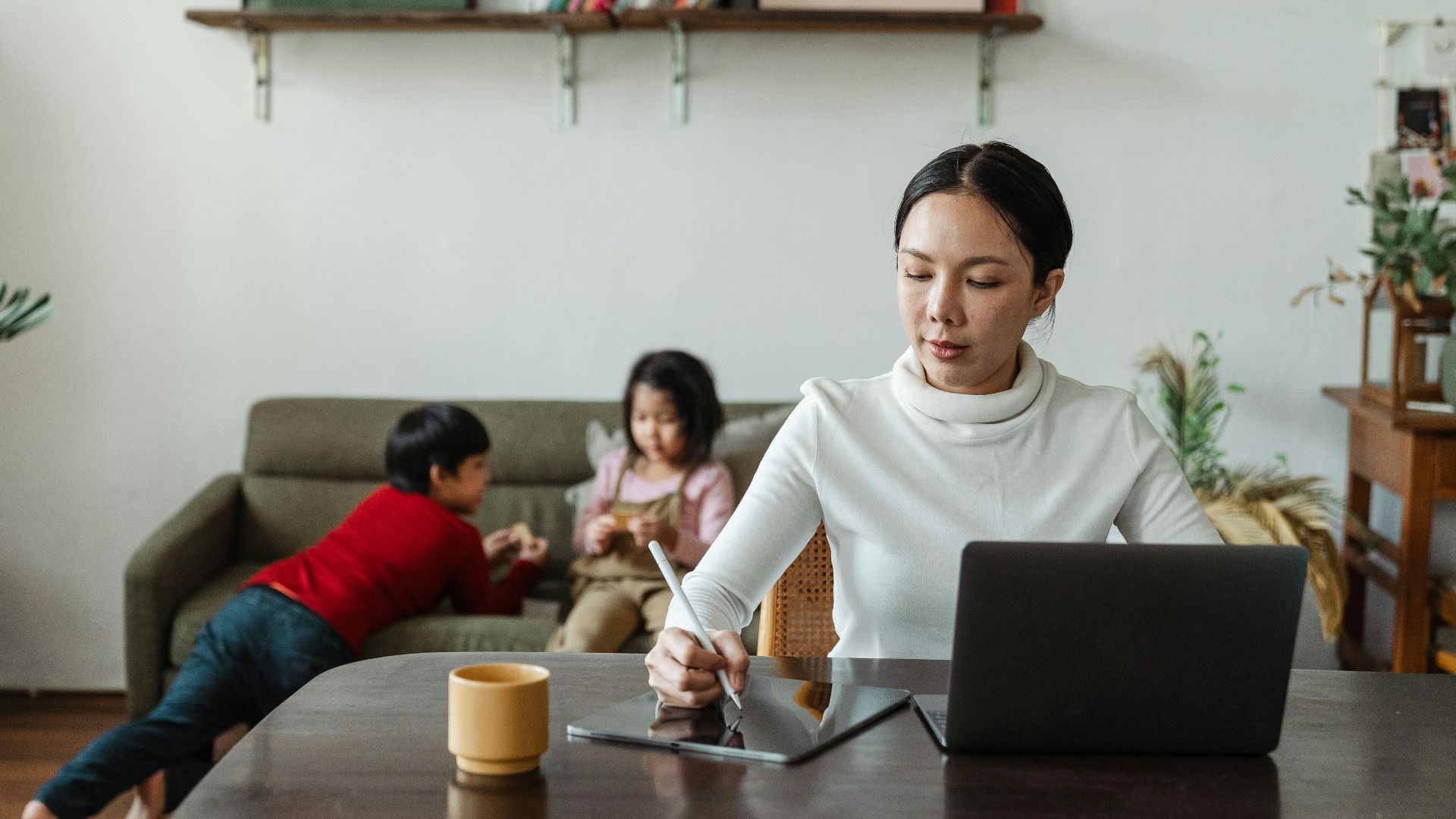 Concentrated young ethnic female using tablet with stylus and laptop during remote work at home with playing children on background