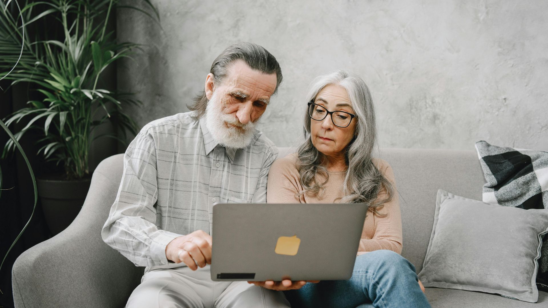 Senior couple sitting on a sofa using a laptop indoors, embracing technology together.
