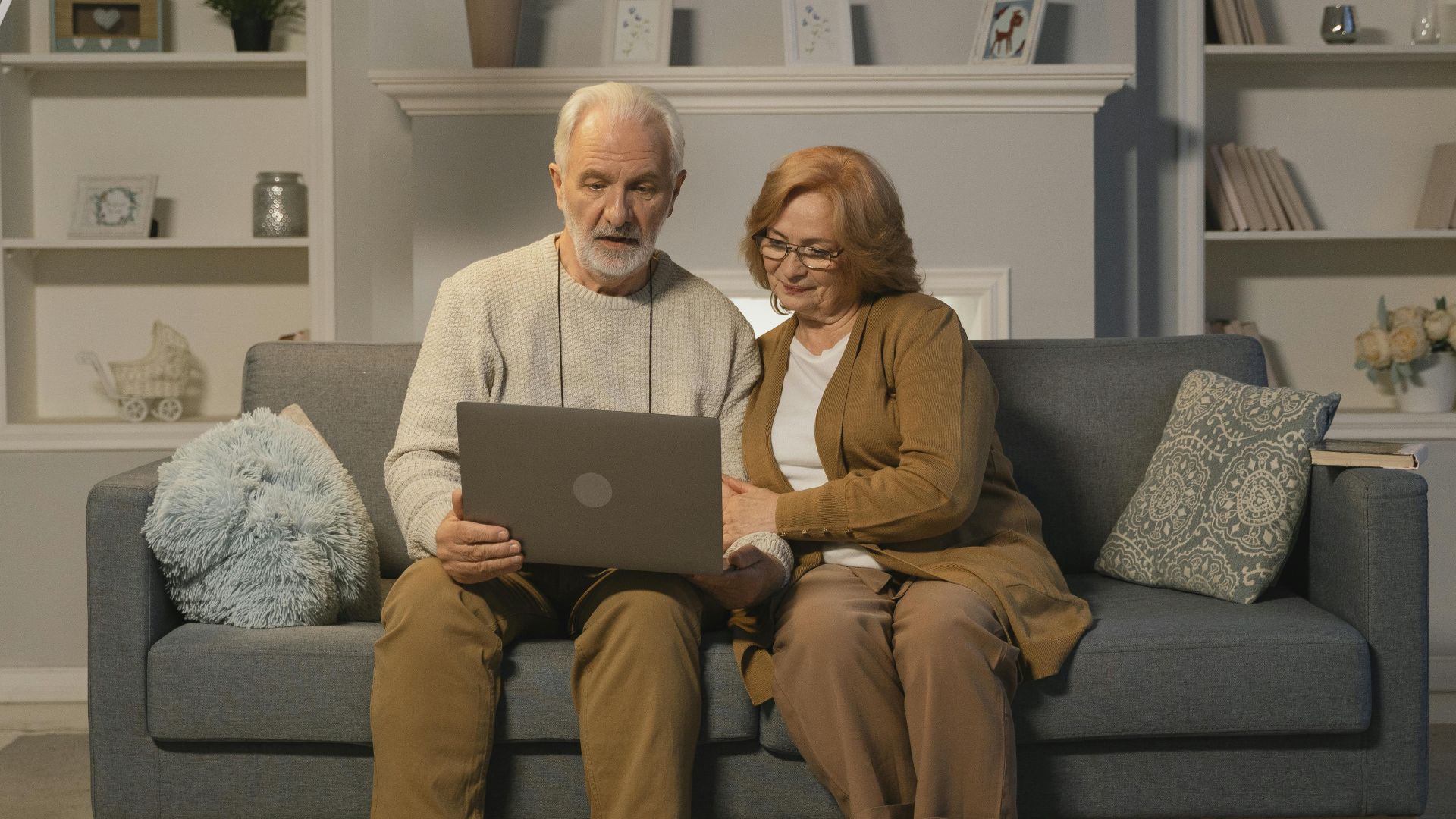 Senior couple sitting on a sofa using a laptop for an online call, surrounded by cozy home decor.