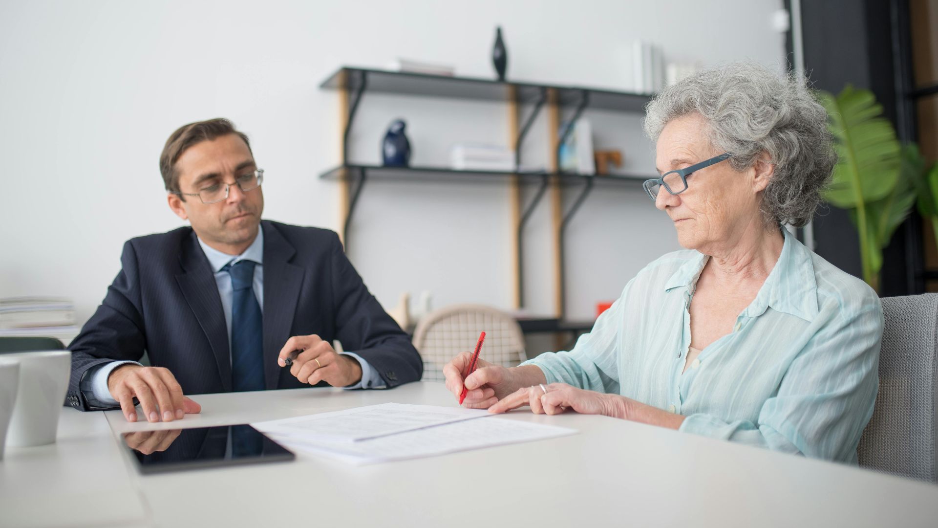 Elderly woman signing paperwork in modern office with consultant.