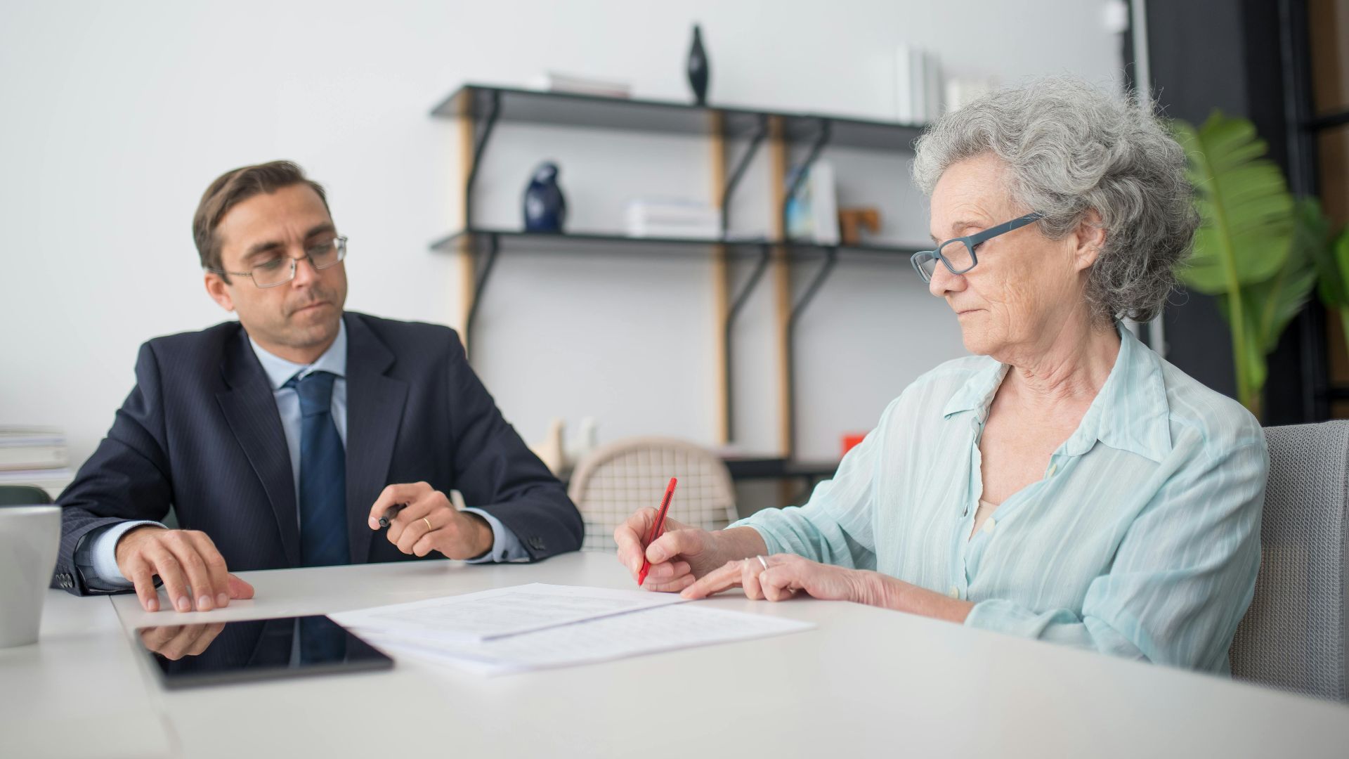 Elderly woman signing paperwork in modern office with consultant.