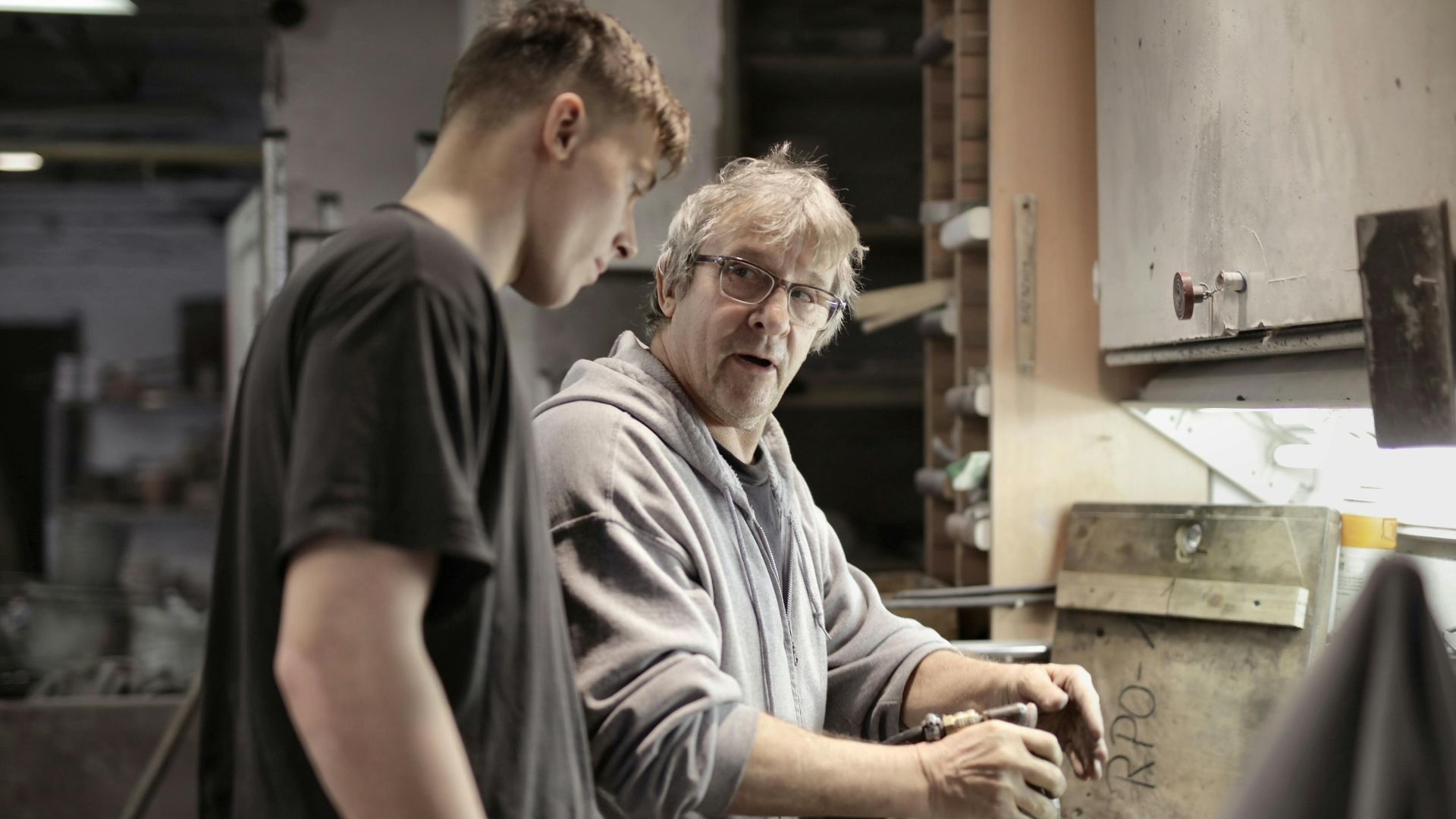 Side view of senior foreman in eyeglasses showing to trainee how handling detail in workshop