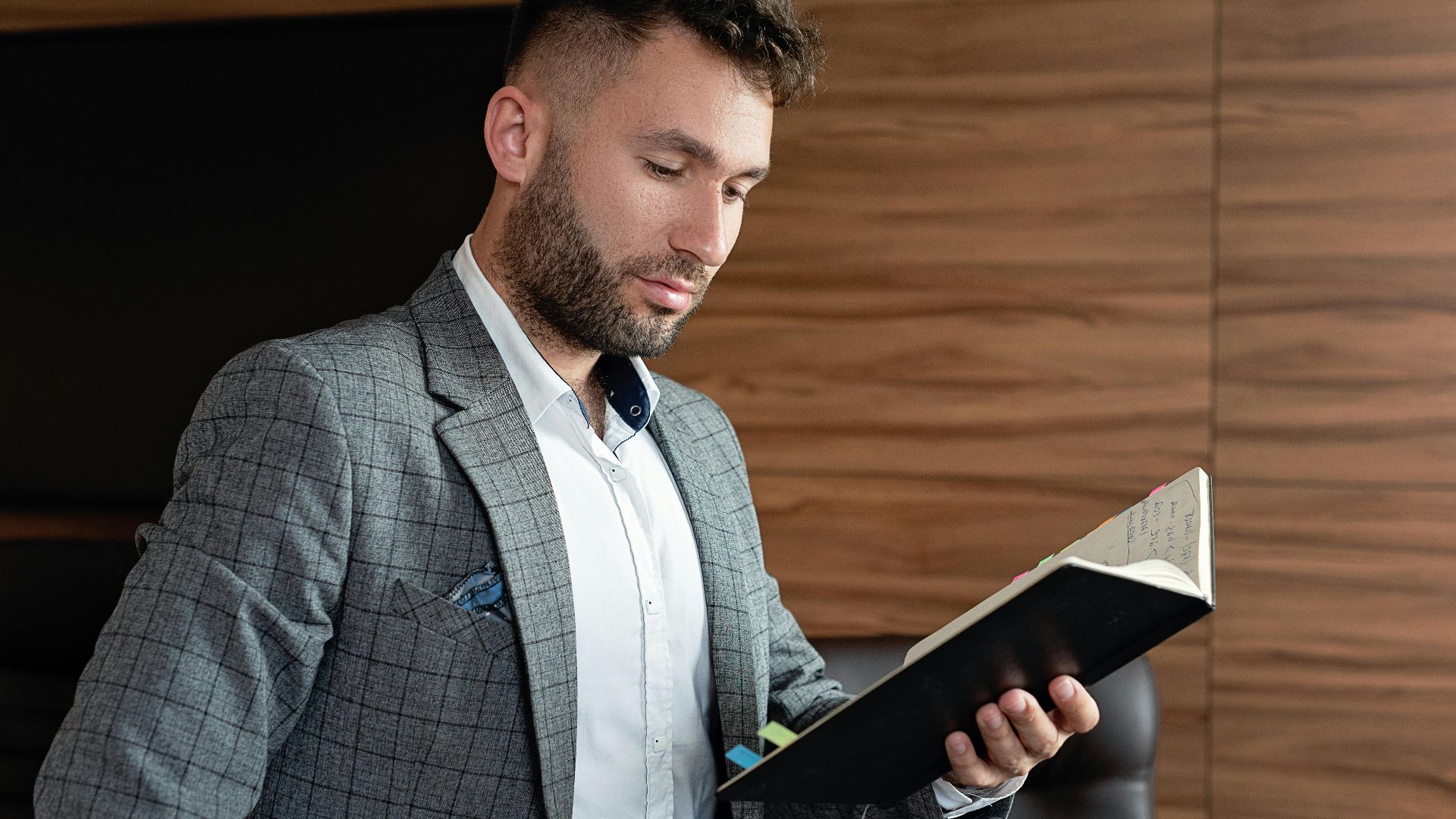 Professional businessman in office setting reading a notebook, dressed in a gray plaid blazer.