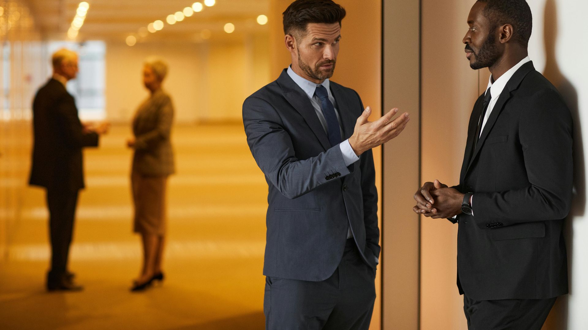 Two businessmen in formal attire engaged in a serious discussion in a modern office hallway.