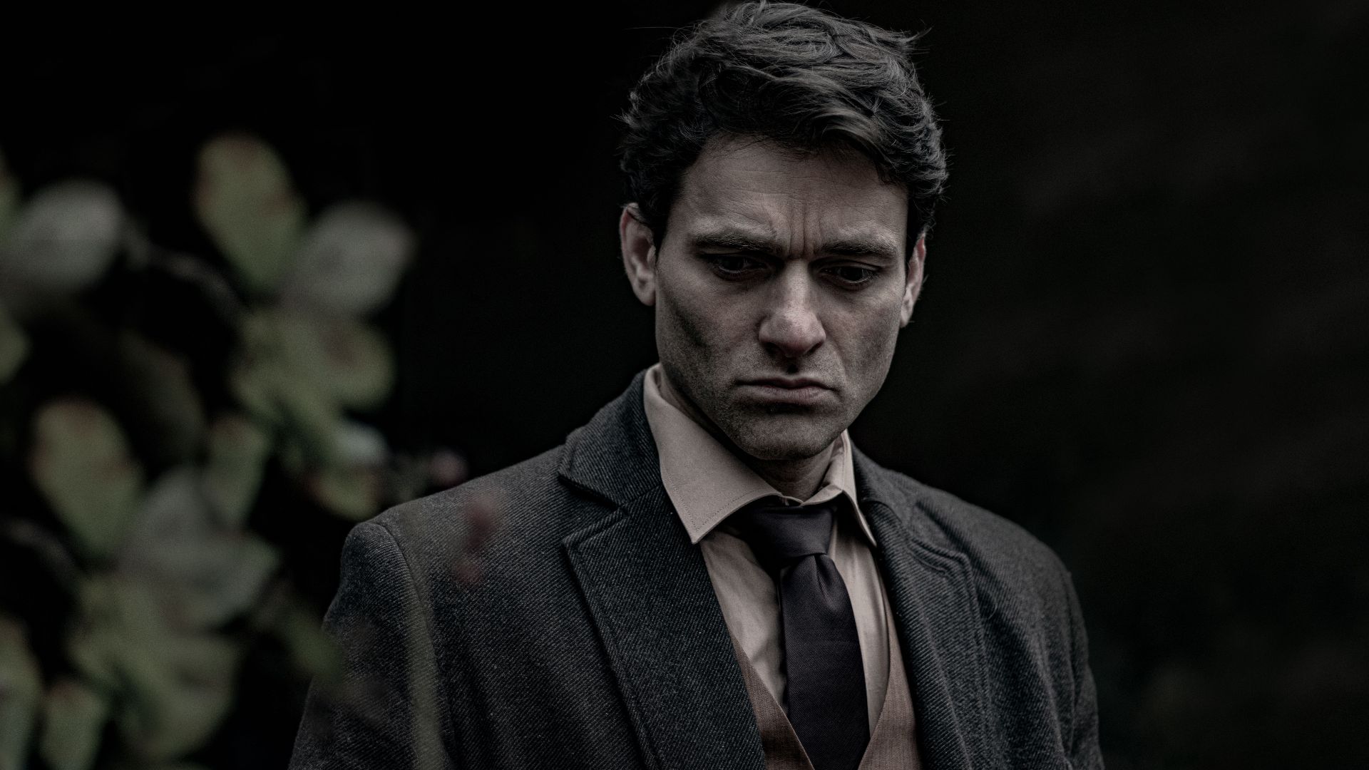 A pensive man stands outdoors in a dark suit, reflecting somberly.