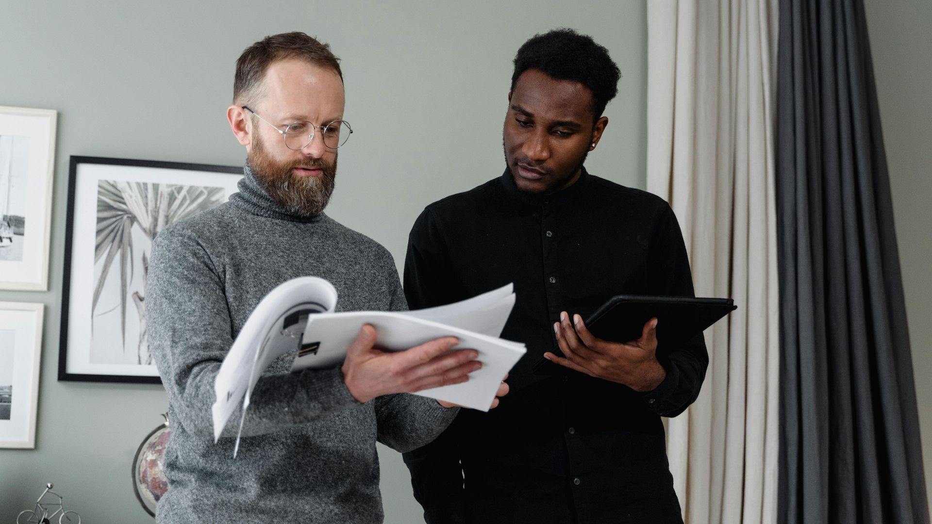 Two businessmen analyzing documents and using a tablet in a modern office setting.