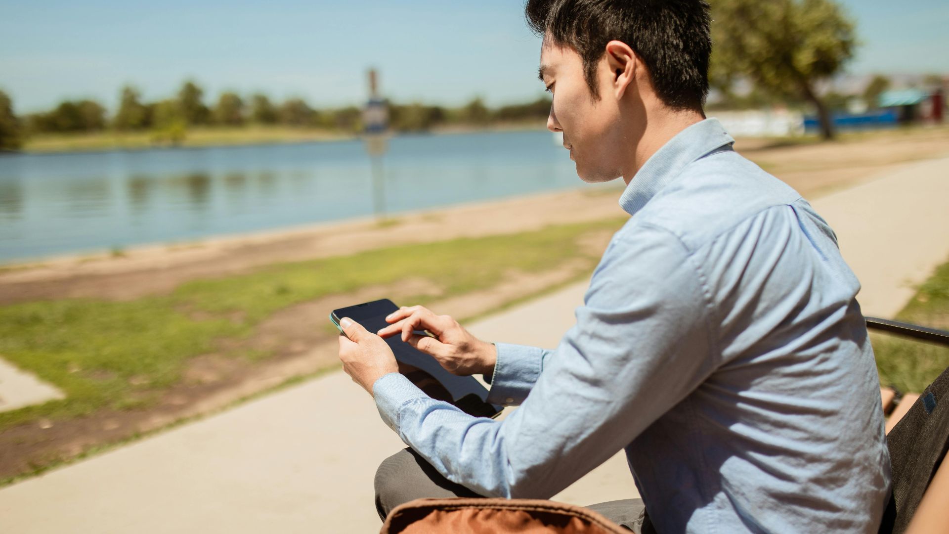 Asian man in blue shirt using a digital tablet while sitting on a riverside bench outdoors.