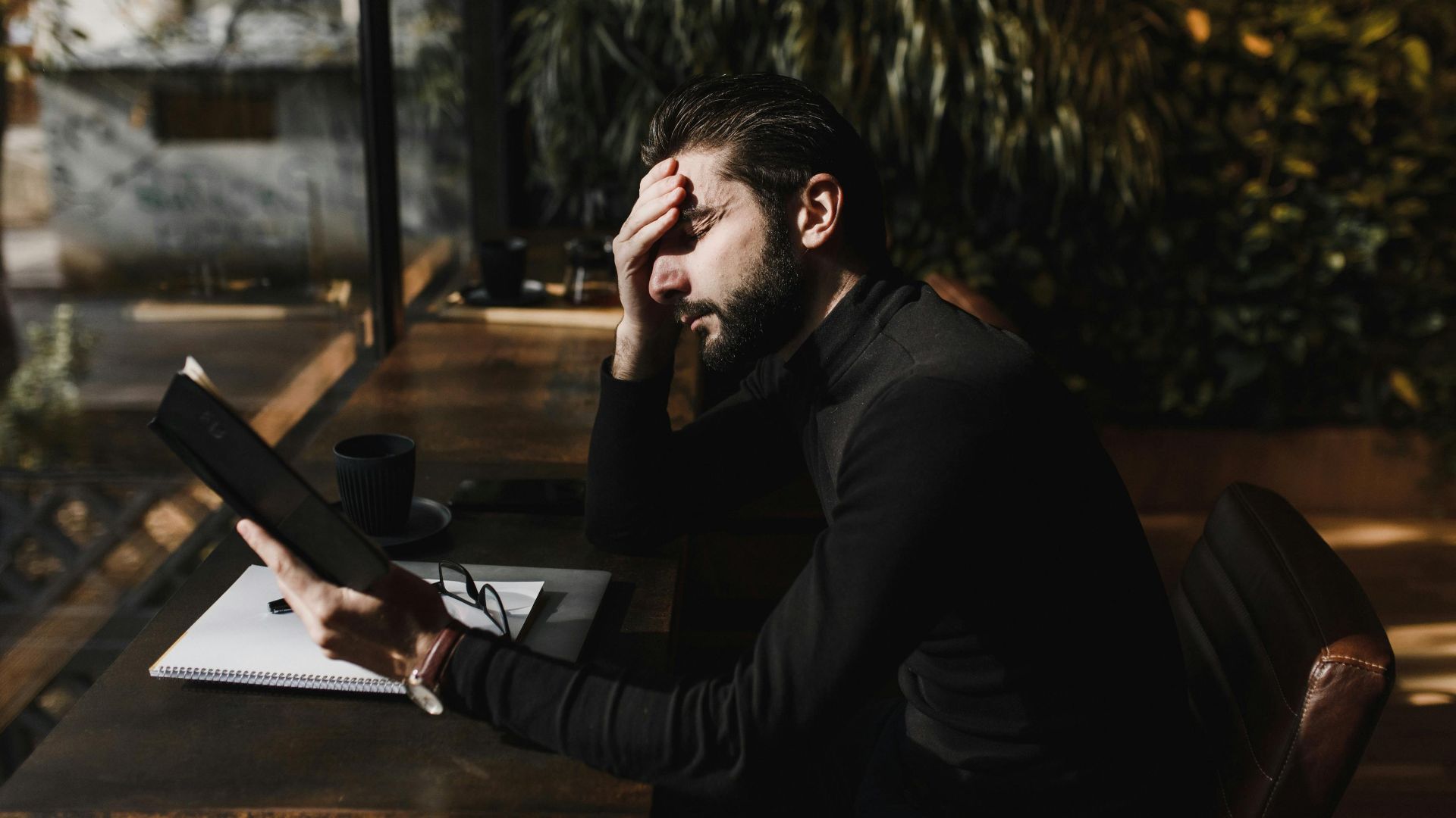 A stressed man with hand on forehead reads in a sunlit room, illustrating concentration and pressure.