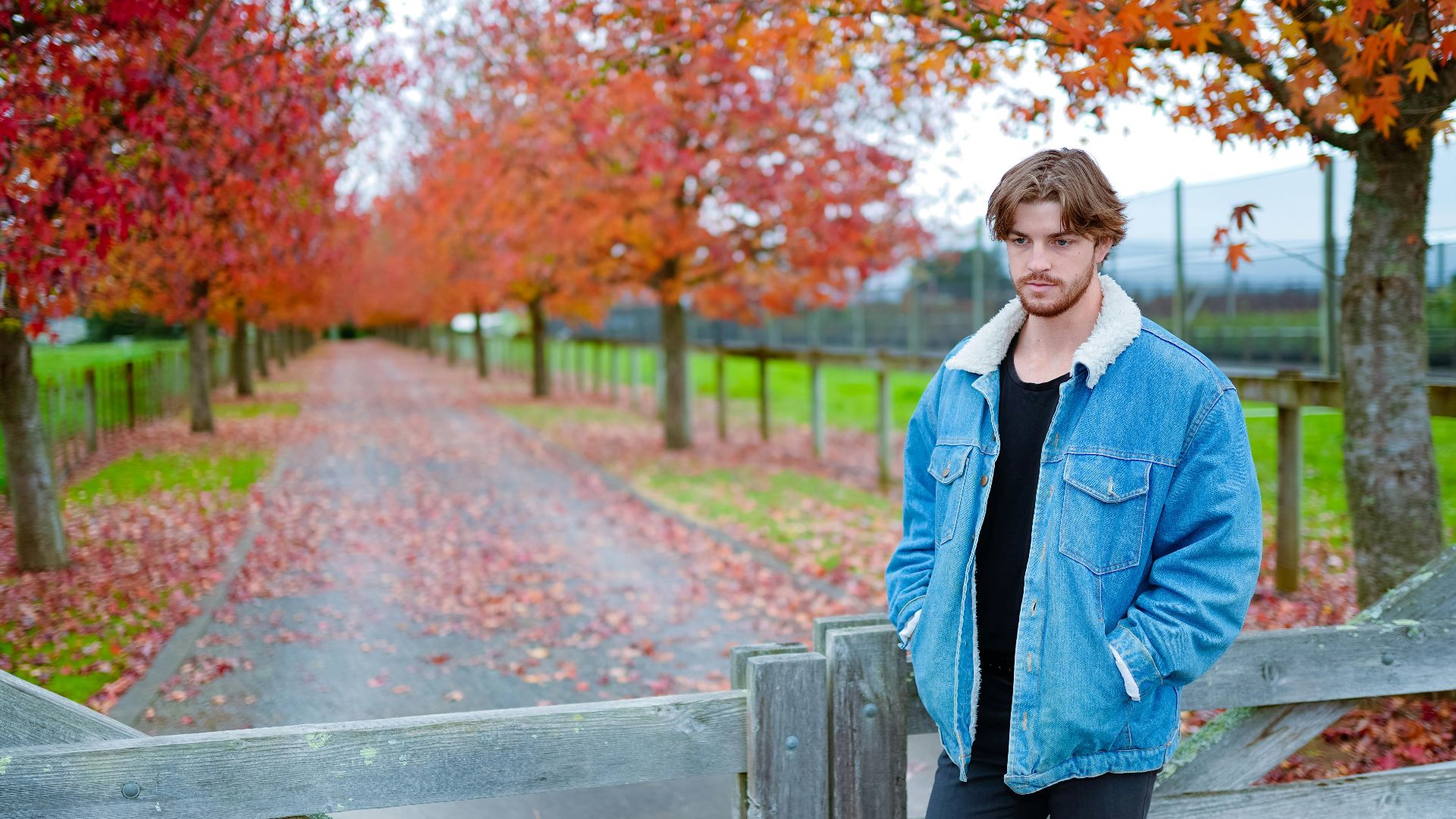 Young man in a denim jacket posing outdoors amidst fall foliage.