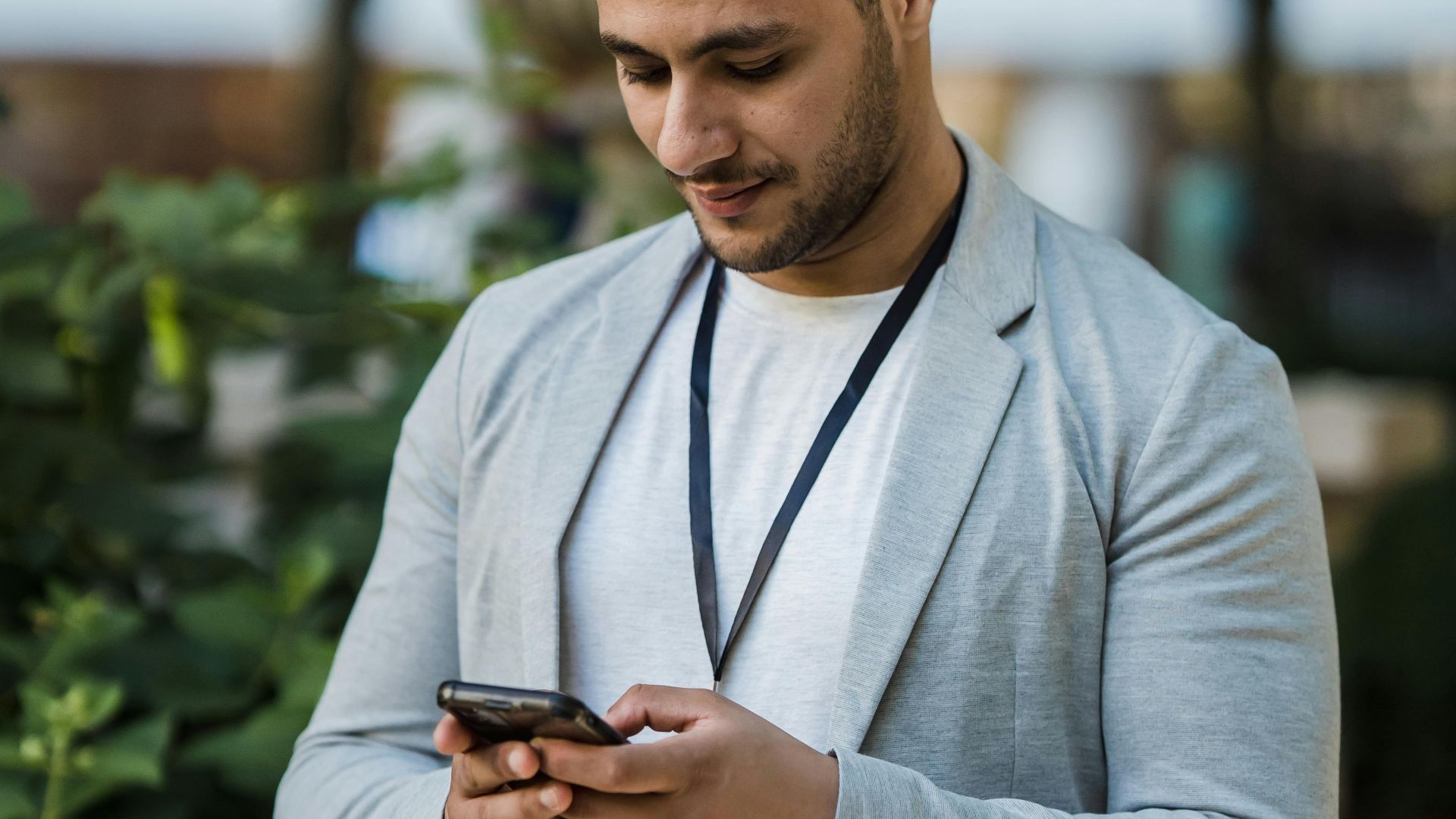 Smiling man using smartphone outdoors in a city park, enjoying technology and nature.