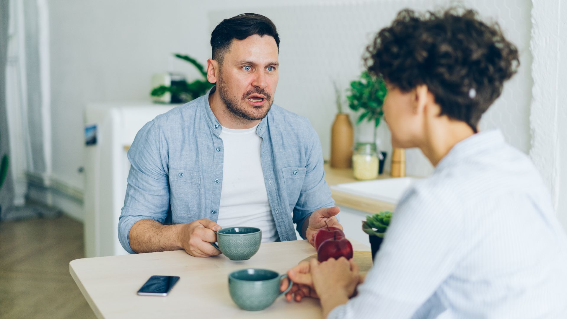 Couple having a serious conversation at the kitchen table with coffee cups.