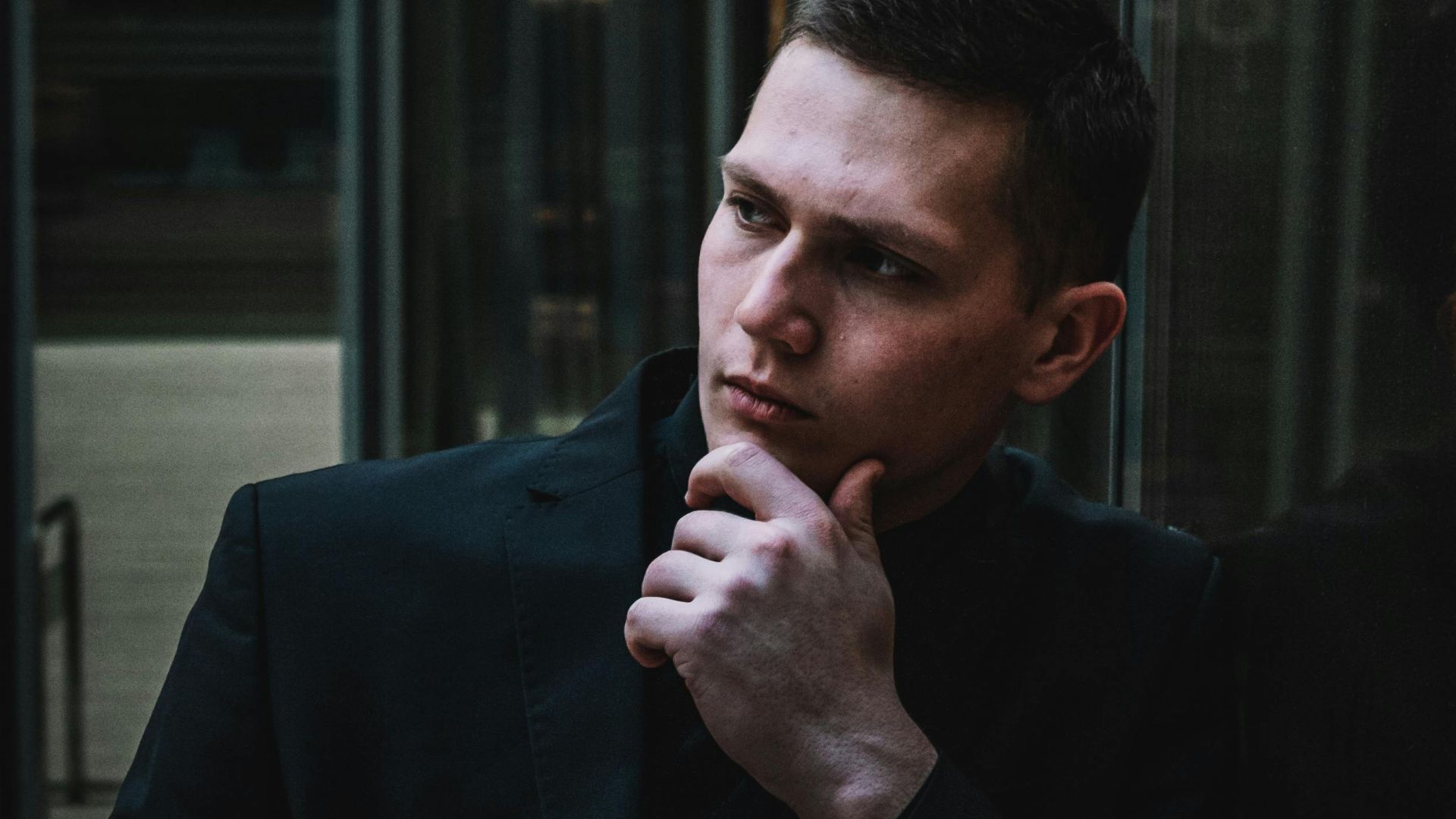 Contemplative man in suit jacket leans against glass wall, reflecting in urban environment.
