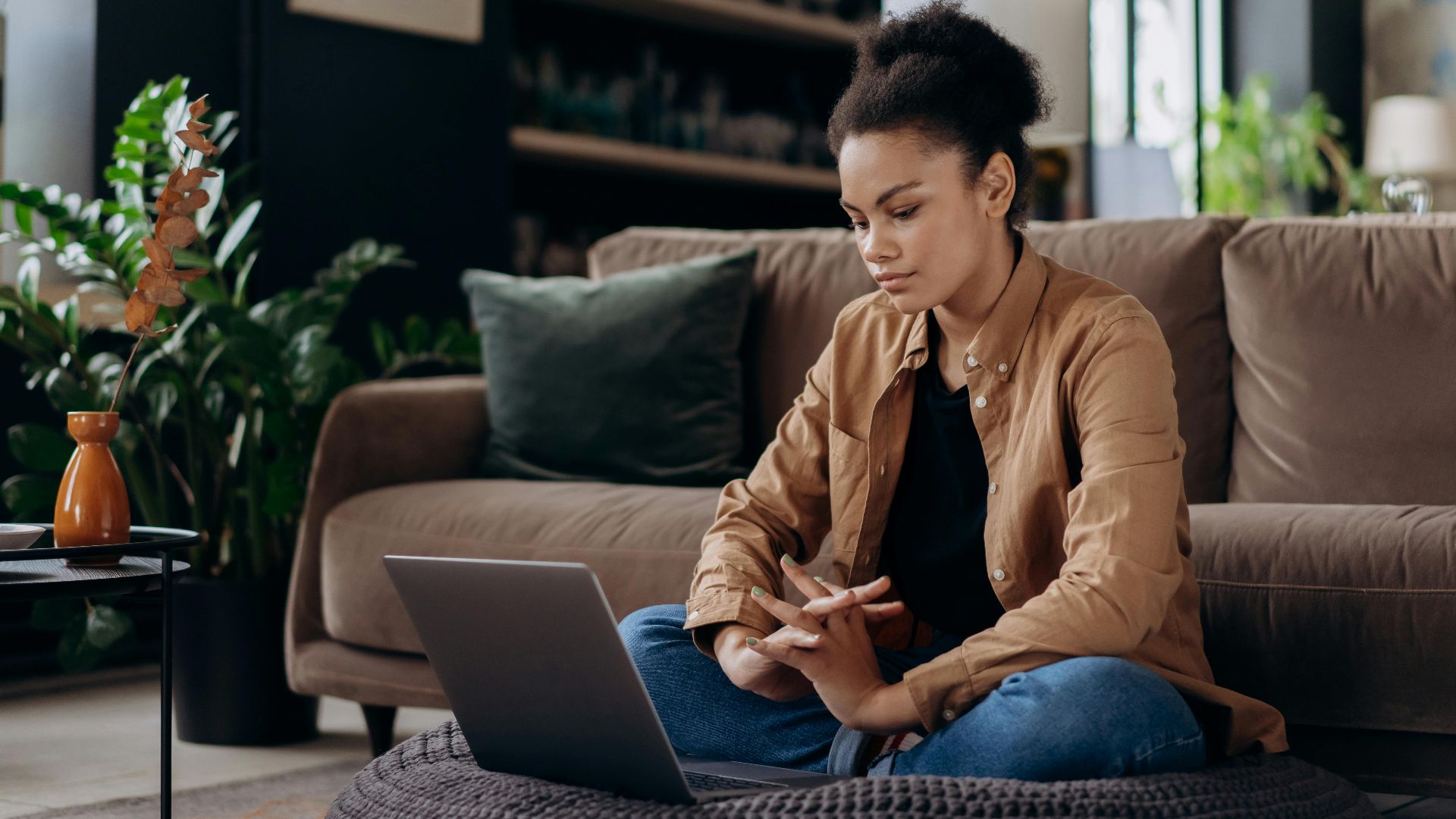 Young woman with afro hair sits on bean bag working on laptop from home, in a cozy living room setting.