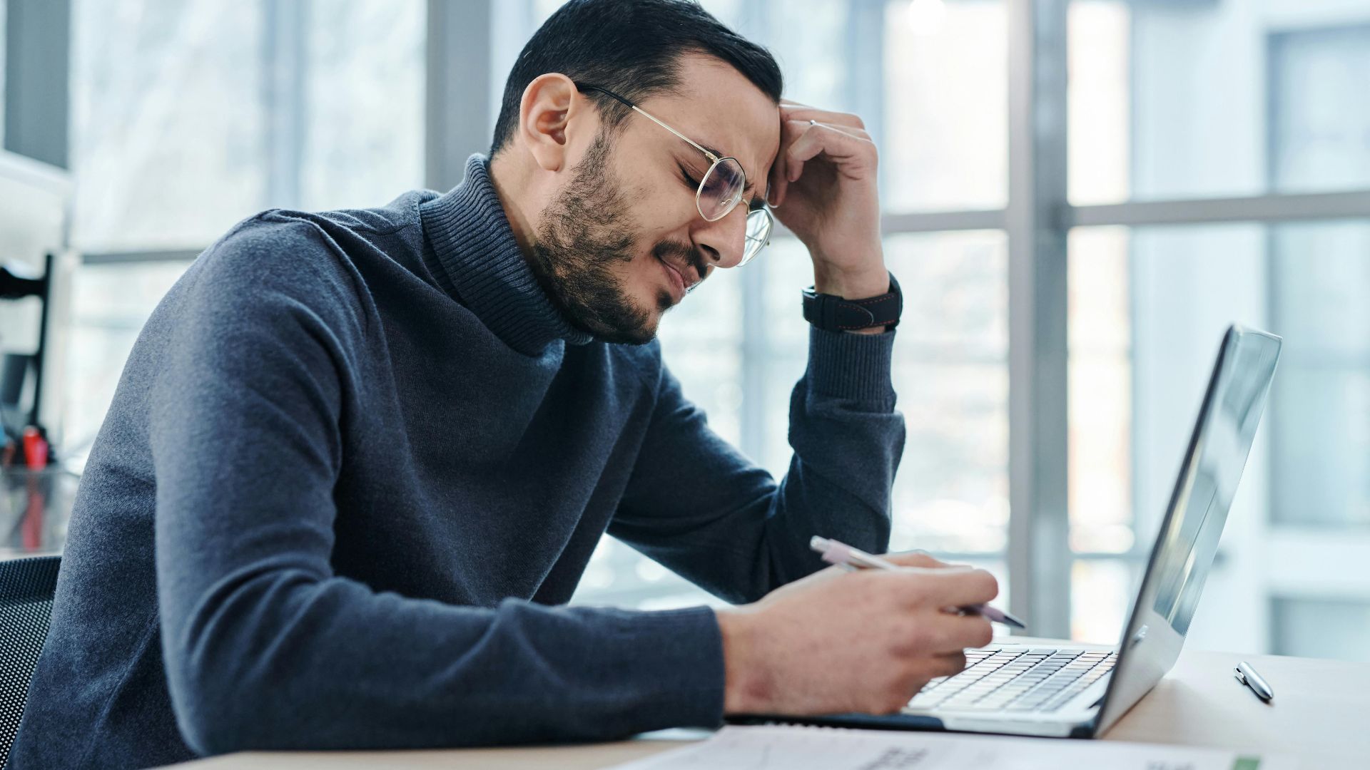 Man looking stressed at laptop in a modern office with large windows.