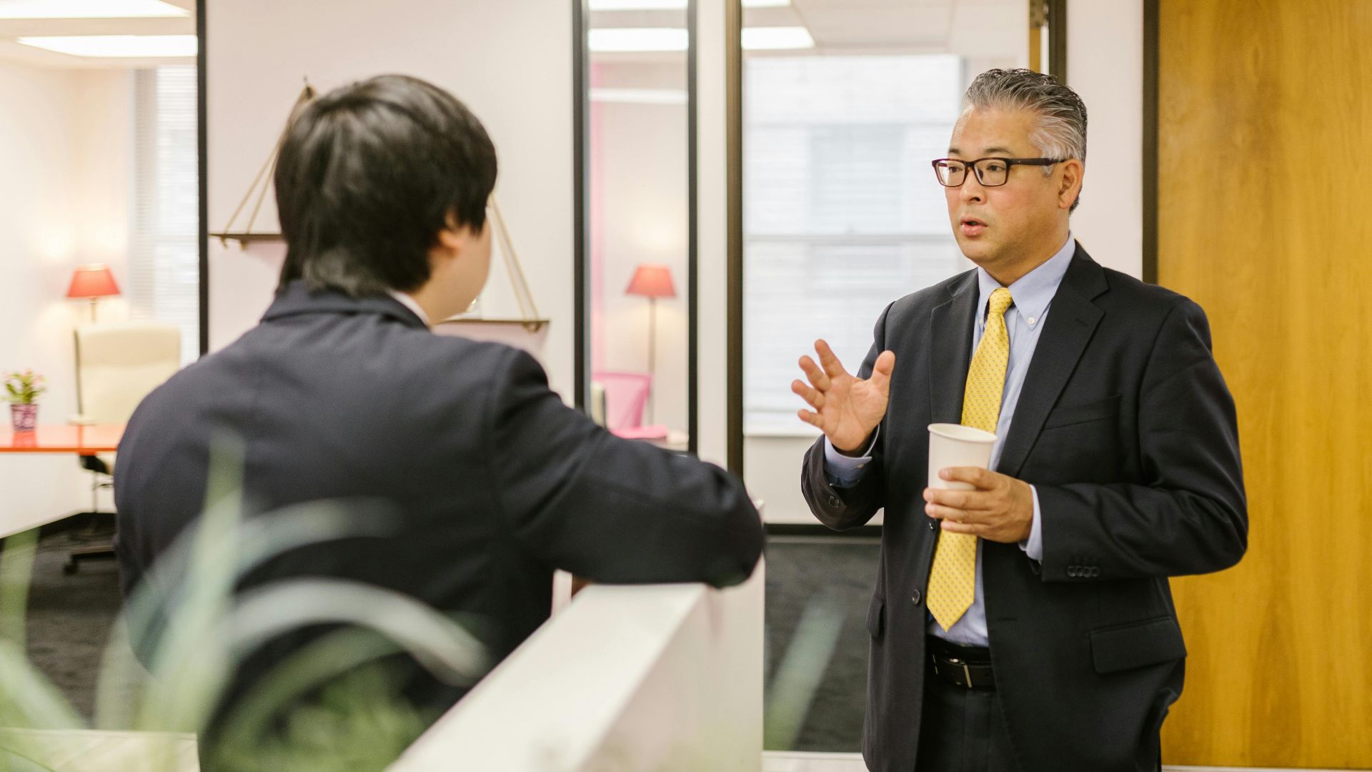 Two businessmen in formal attire having a conversation in an office setting.