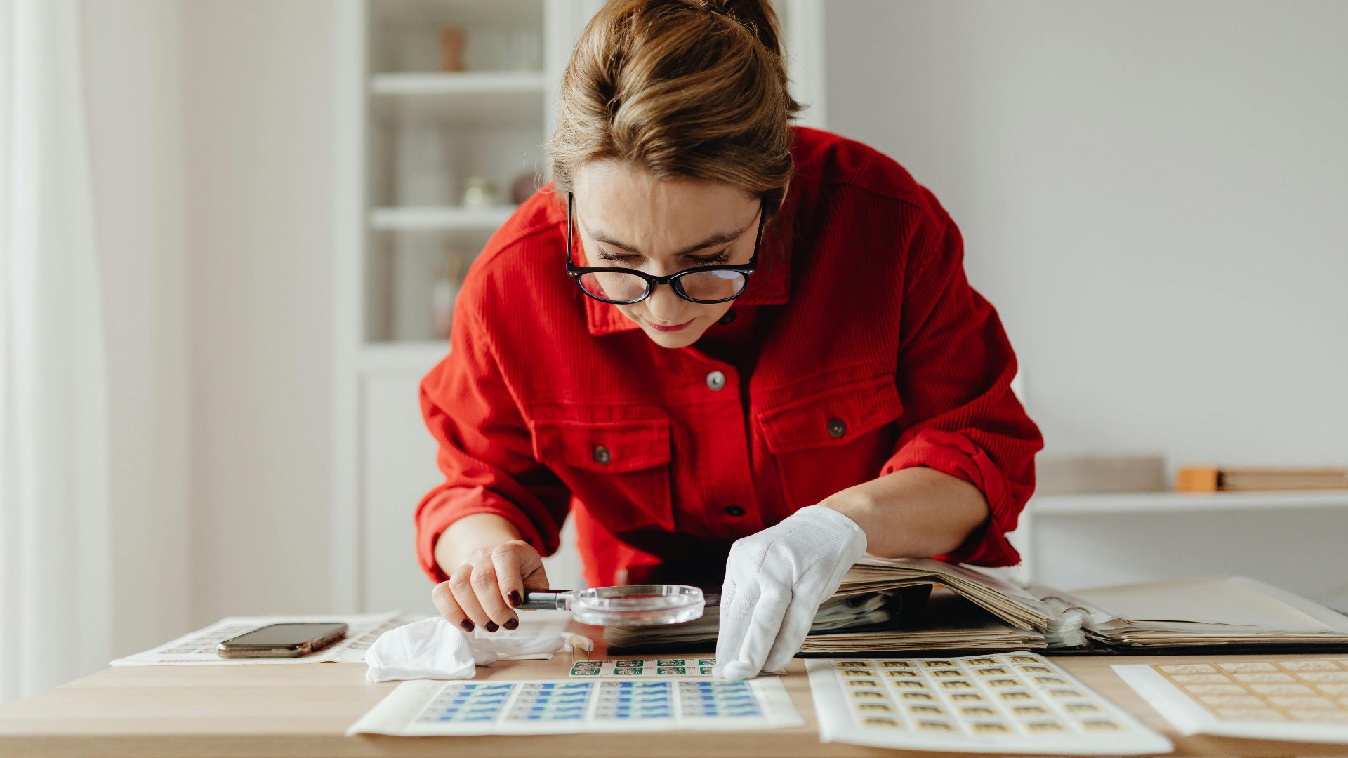 Woman in red shirt examines postage stamps with magnifying glass indoors.