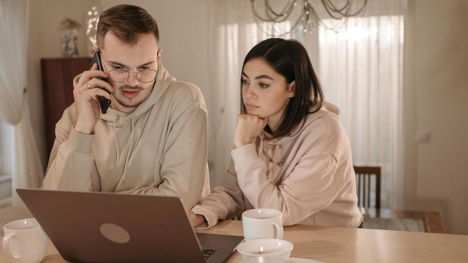 A couple in hoodies focused on work using a laptop and smartphone at home.