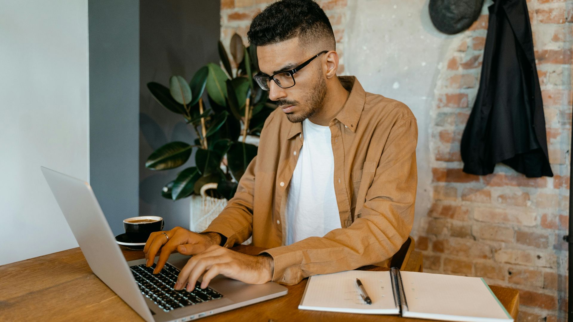 Man in casual office setting typing on laptop with coffee and notebook on desk.