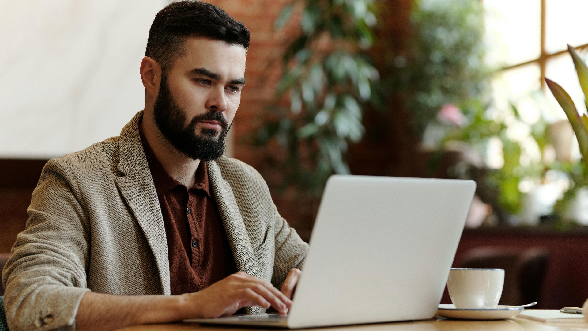 Businessman working on laptop in a cafe setting, surrounded by plants, sipping coffee.