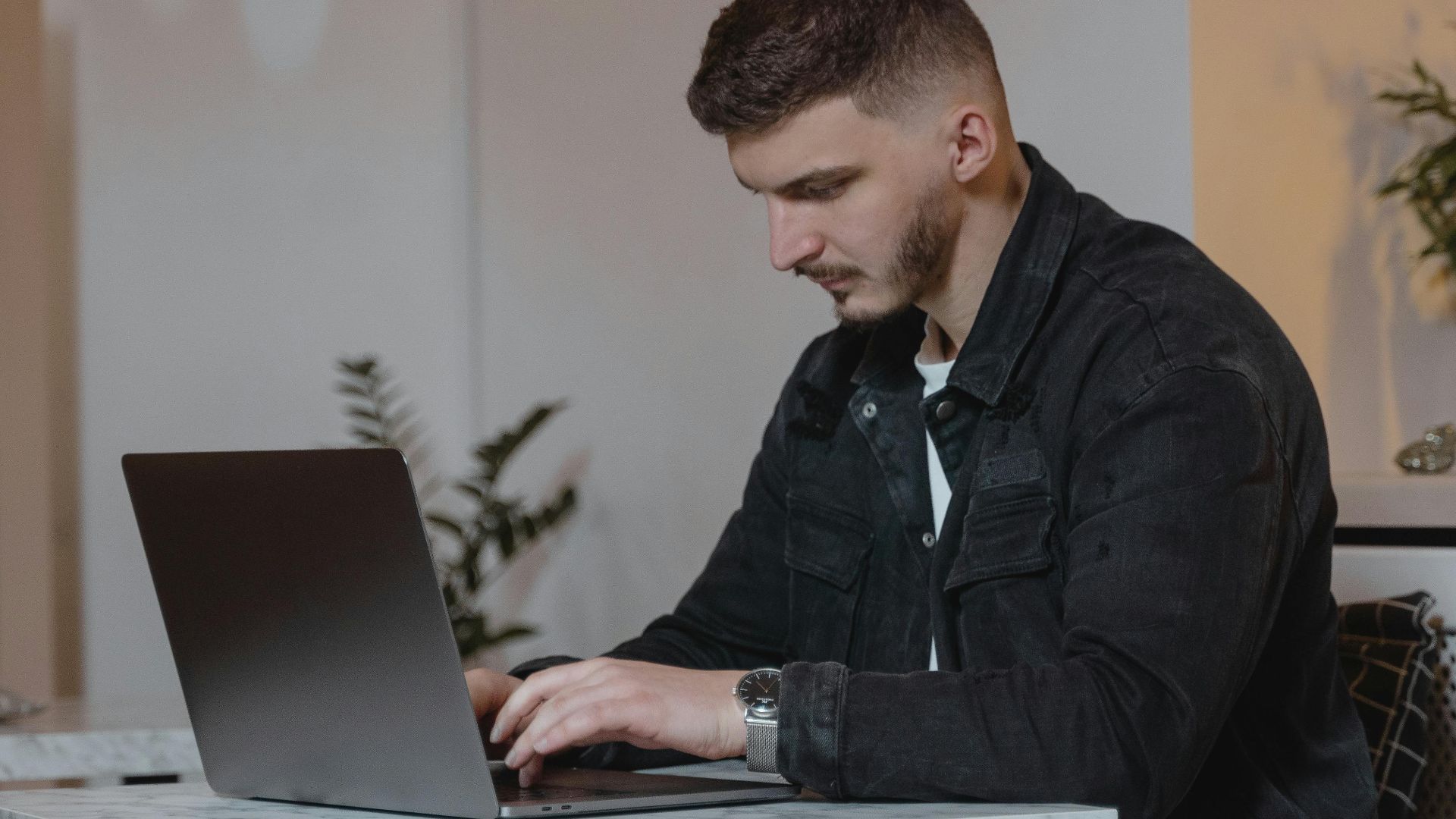 Young man in a denim jacket working on a laptop indoors, showcasing modern remote work lifestyle.