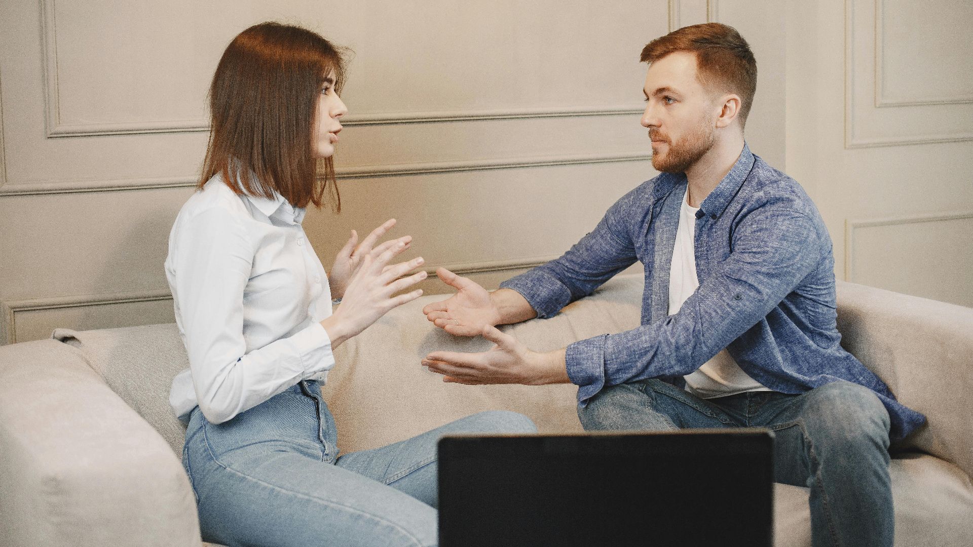 A young couple sitting on a couch having a thoughtful conversation with expressive hand gestures.