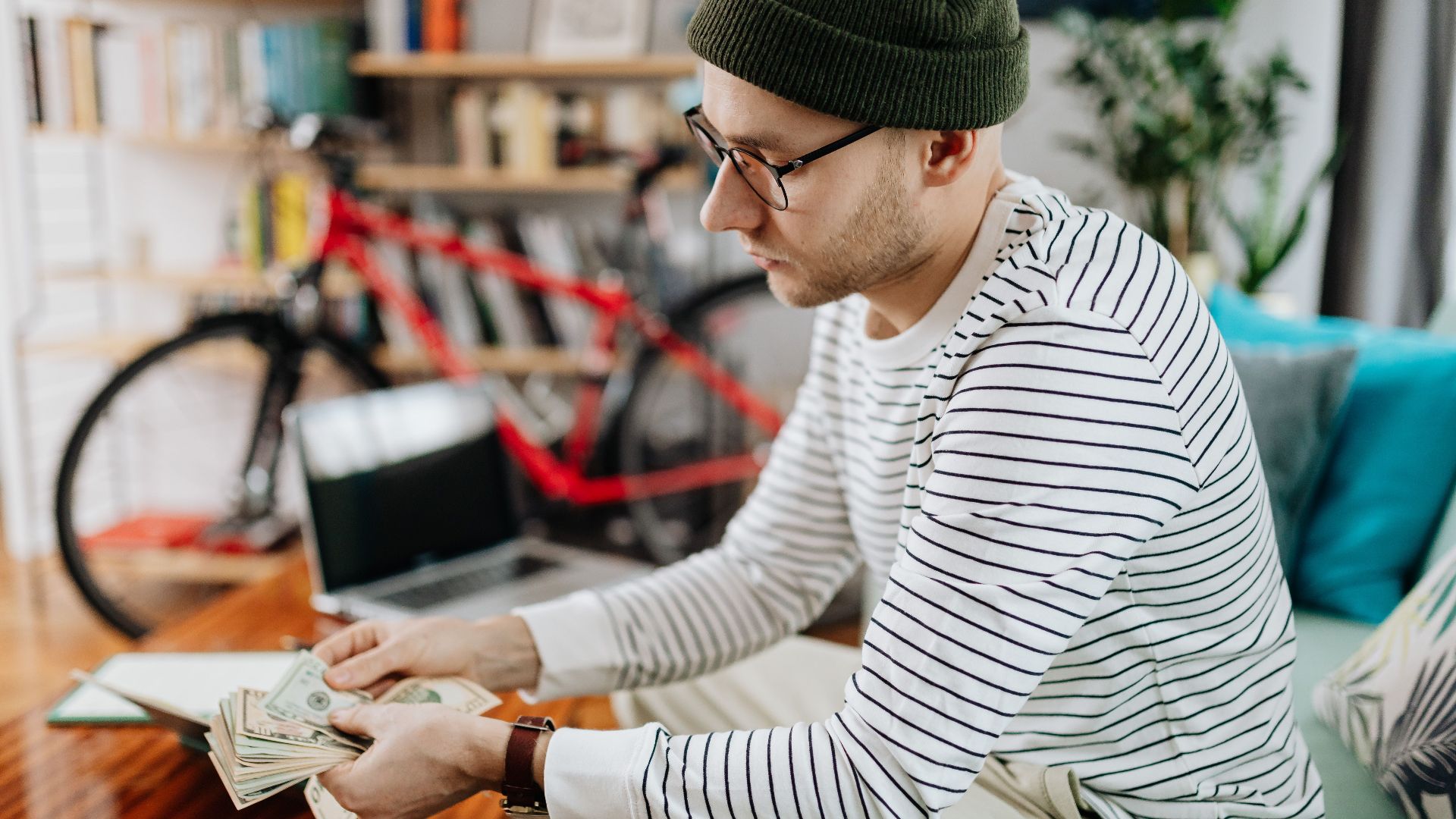 Man in striped shirt counting cash indoors, with a bicycle in the background.