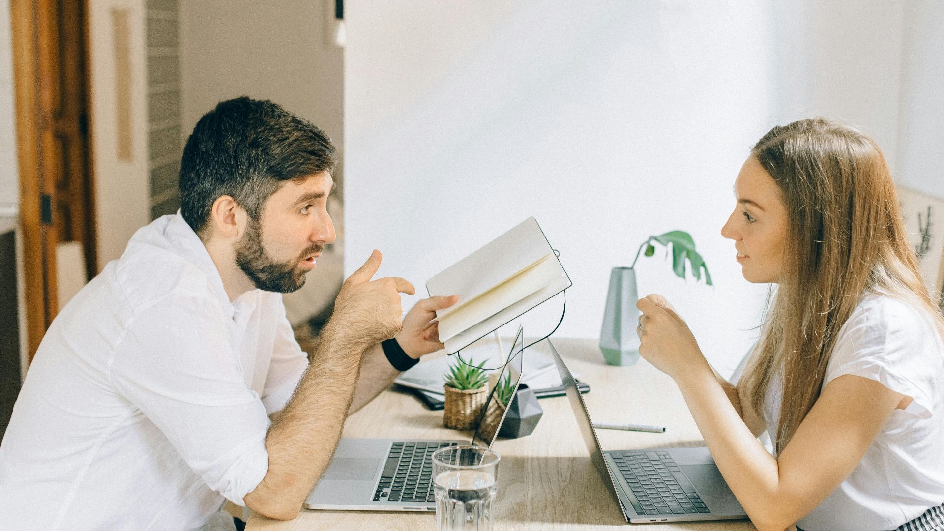 A man and woman working from home on laptops, engaged in discussion at a wooden table.