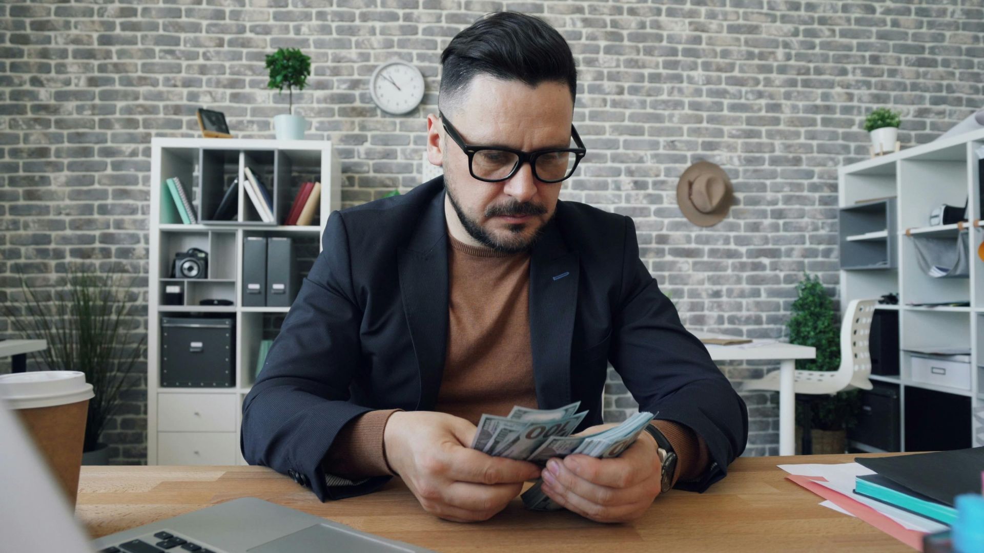 Businessman counting cash at a desk in a modern office setting with a laptop and documents.