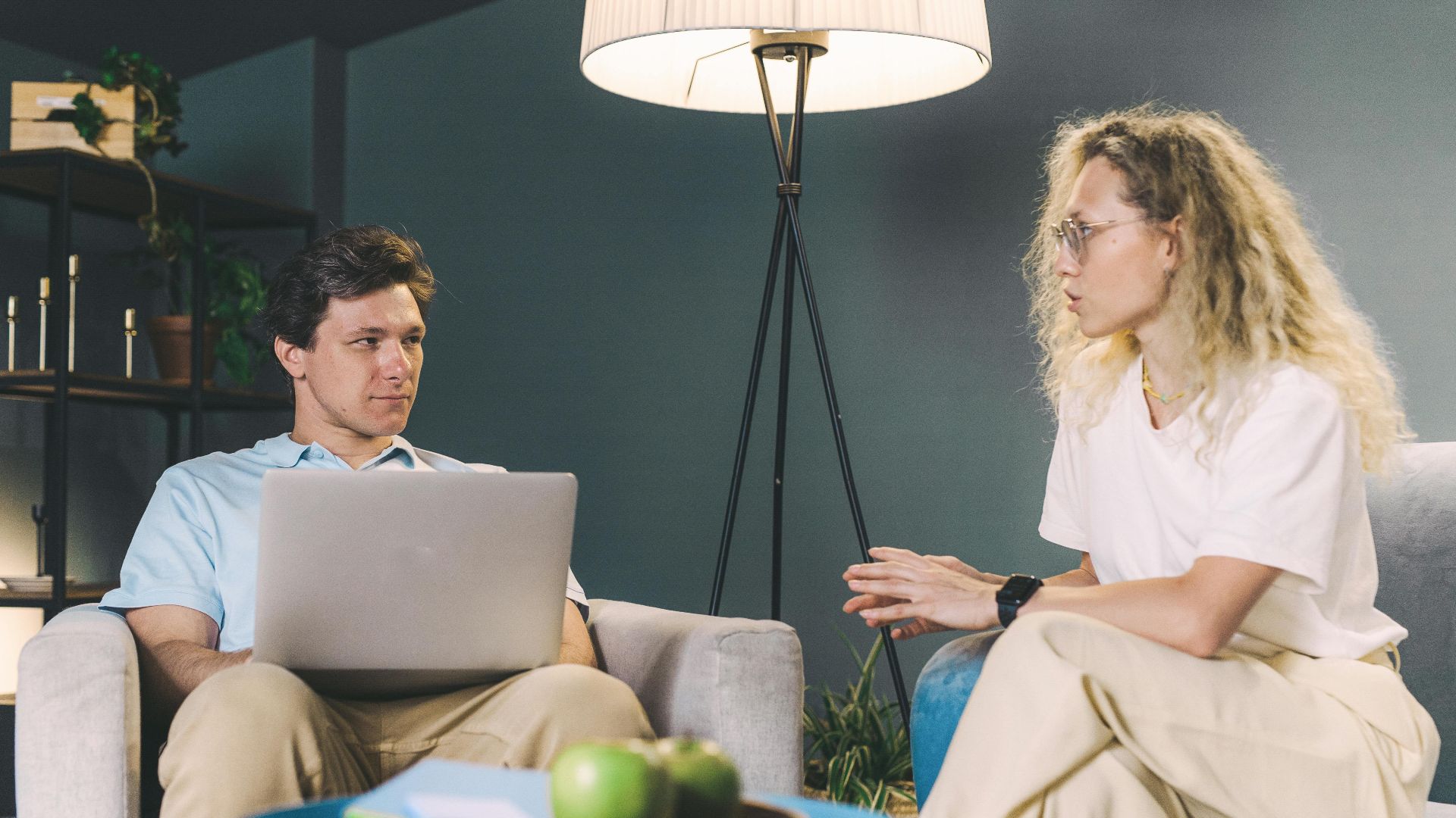 Two adults discuss work while sitting on sofas in a cozy, modern living room setting.