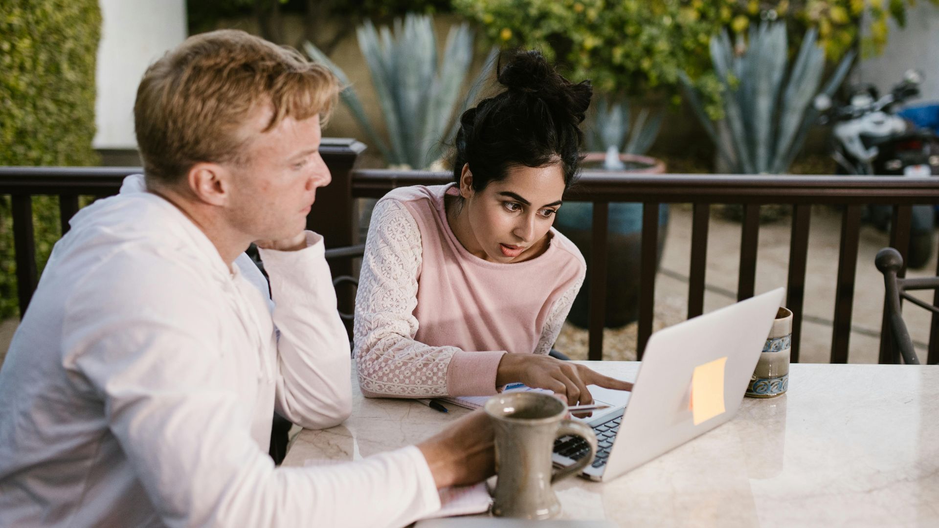 Two adults collaborating on a laptop, seated outdoors with coffee on the table.